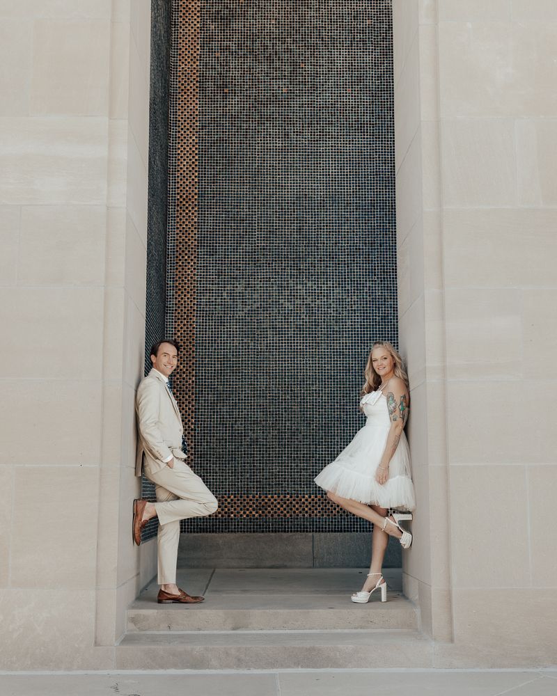A couple in white outfits poses playfully in an architectural alcove with a dark textured wall panel.