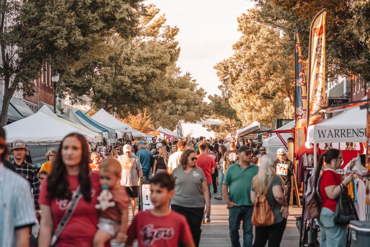 Crowded outdoor street festival with vendor tents, tall trees, and people walking under warm golden sunlight.