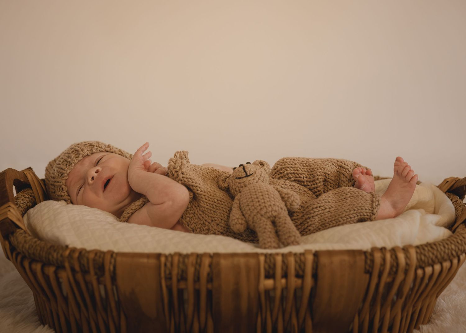 Newborn baby in knit outfit sleeping in wicker basket with teddy bear toy on white fur blanket.