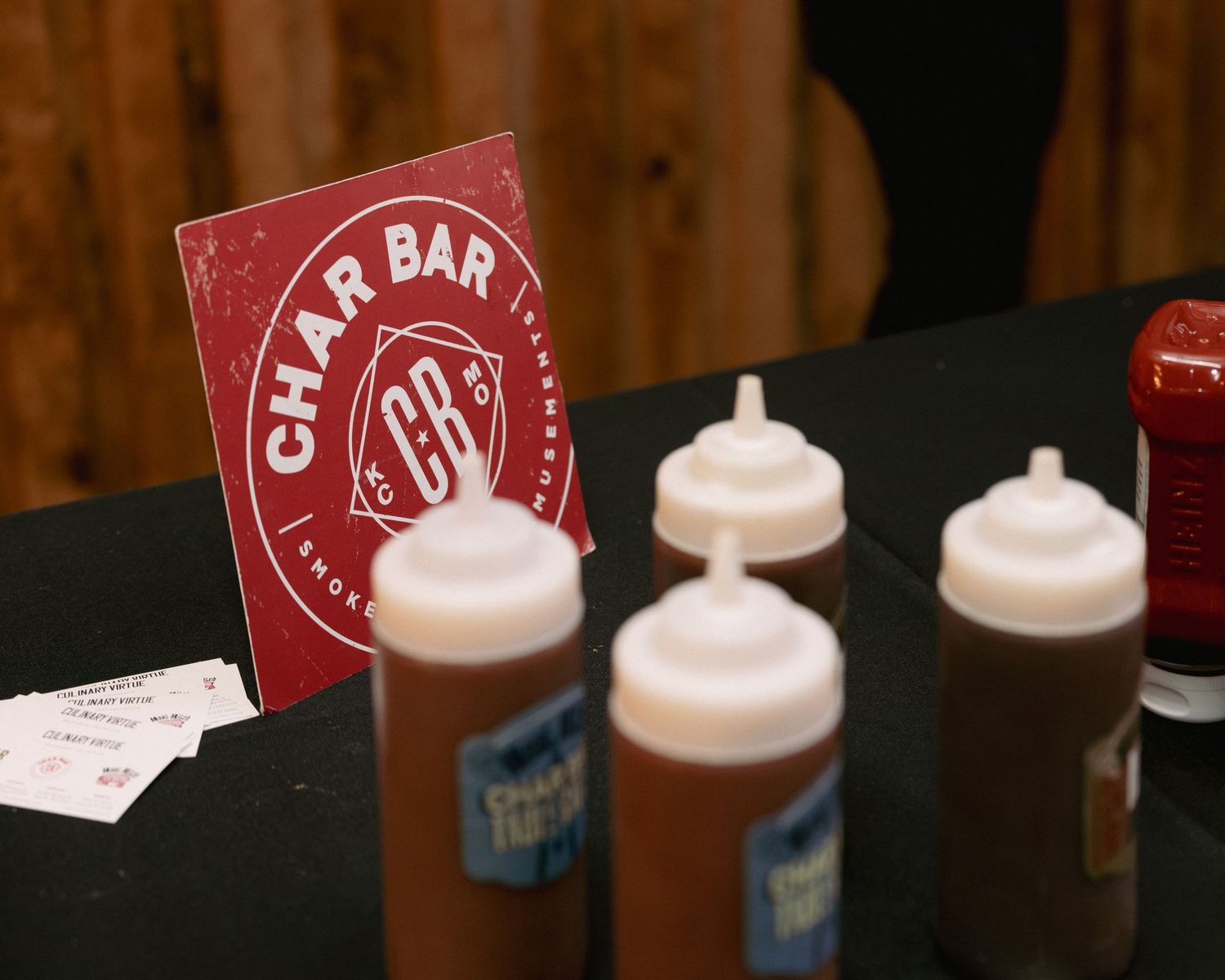 Char Bar BBQ sauce bottles and menu sign displayed on a black table at a smoky barbecue restaurant event.