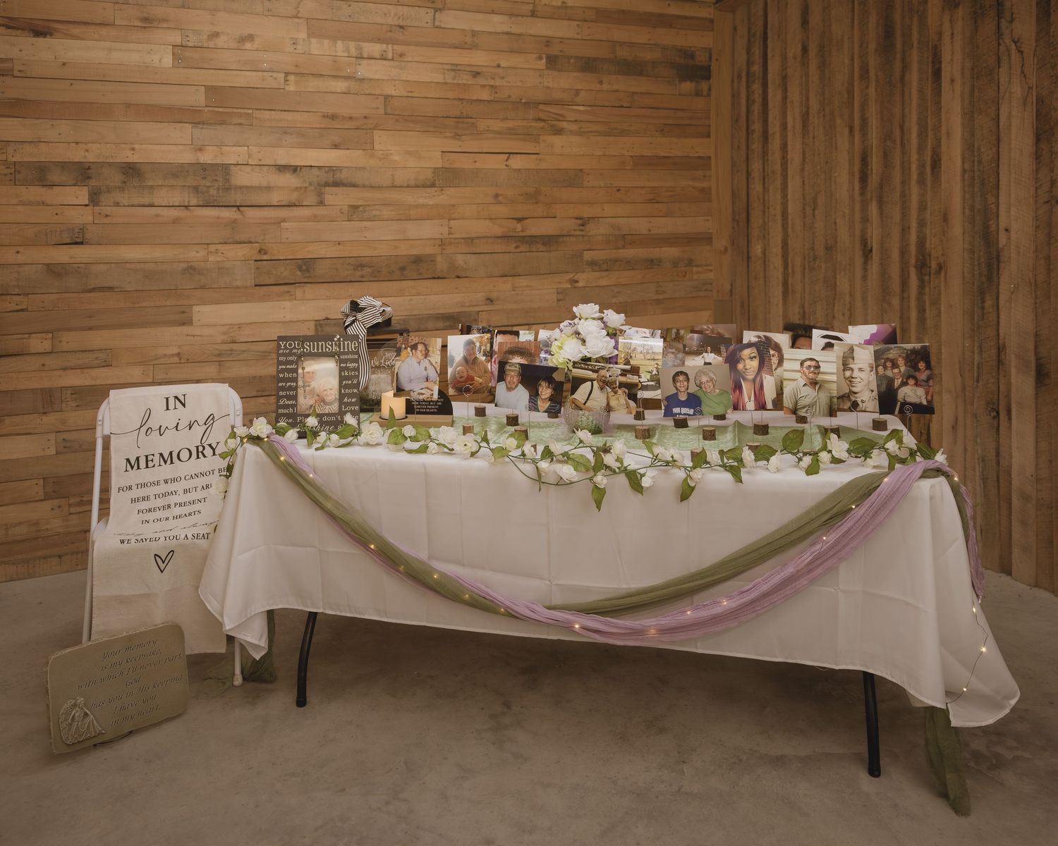 A memorial table with photos, flowers, and decorations displayed on a white tablecloth against a wooden wall.