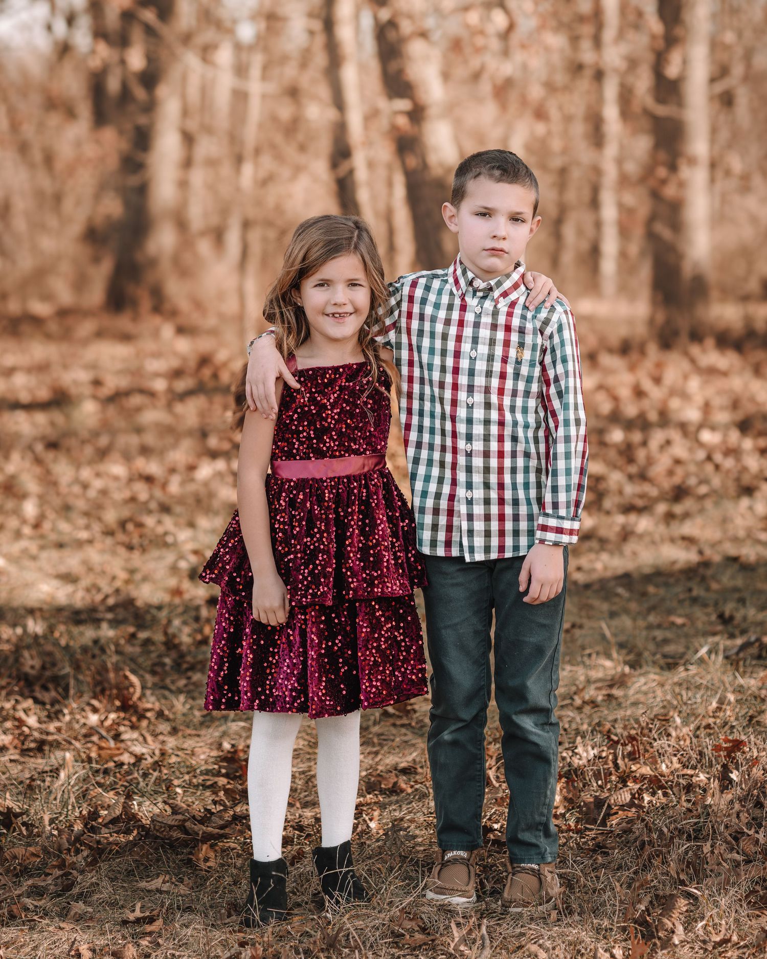 Two children in fall clothing stand together in a wooded area with autumn leaves on the ground.