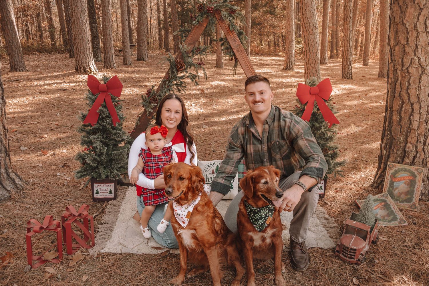 Family and their Irish Setter dog pose for holiday photos in a rustic forest setting decorated with red bows and greenery.