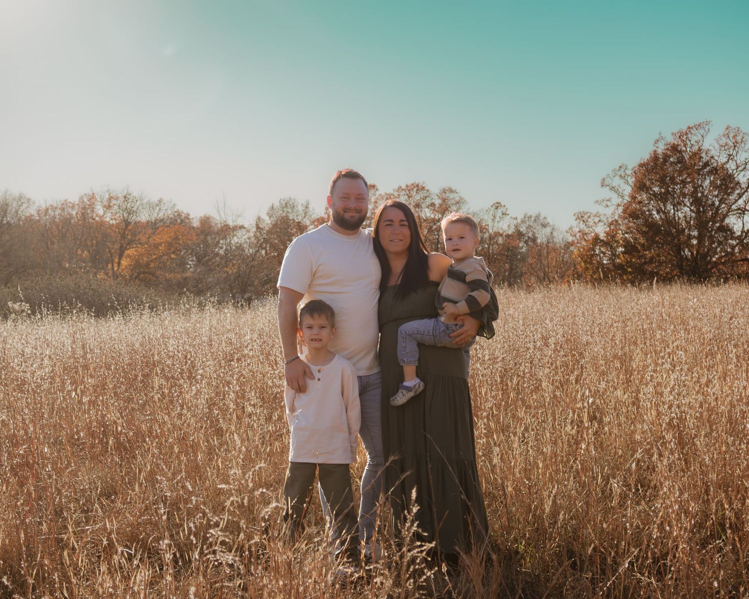 A family stands together in a golden field during sunset with warm lighting and a teal sky in the background.