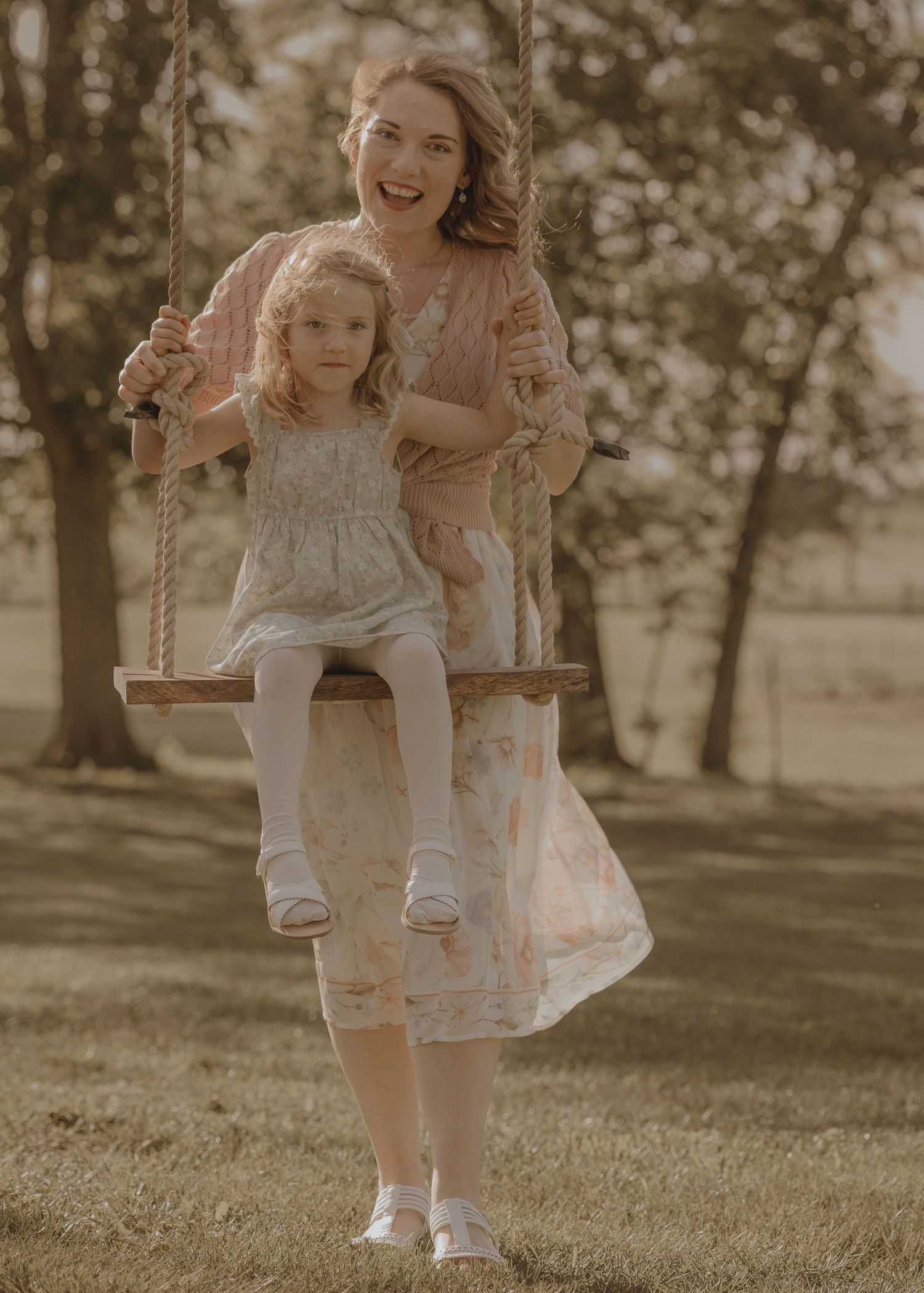A smiling mother in a floral dress pushes her young daughter on a wooden rope swing outdoors.