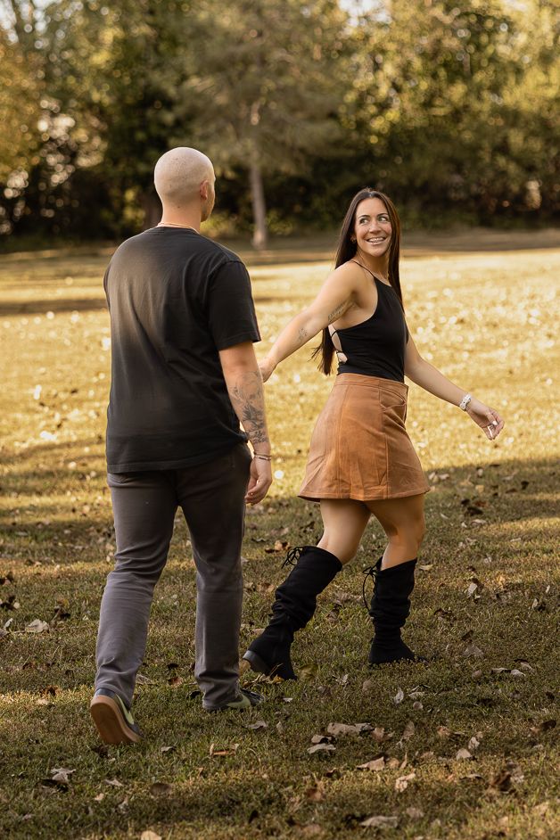 A smiling woman in a tan skirt and boots holds hands with a bald man in a black shirt while walking in a park.