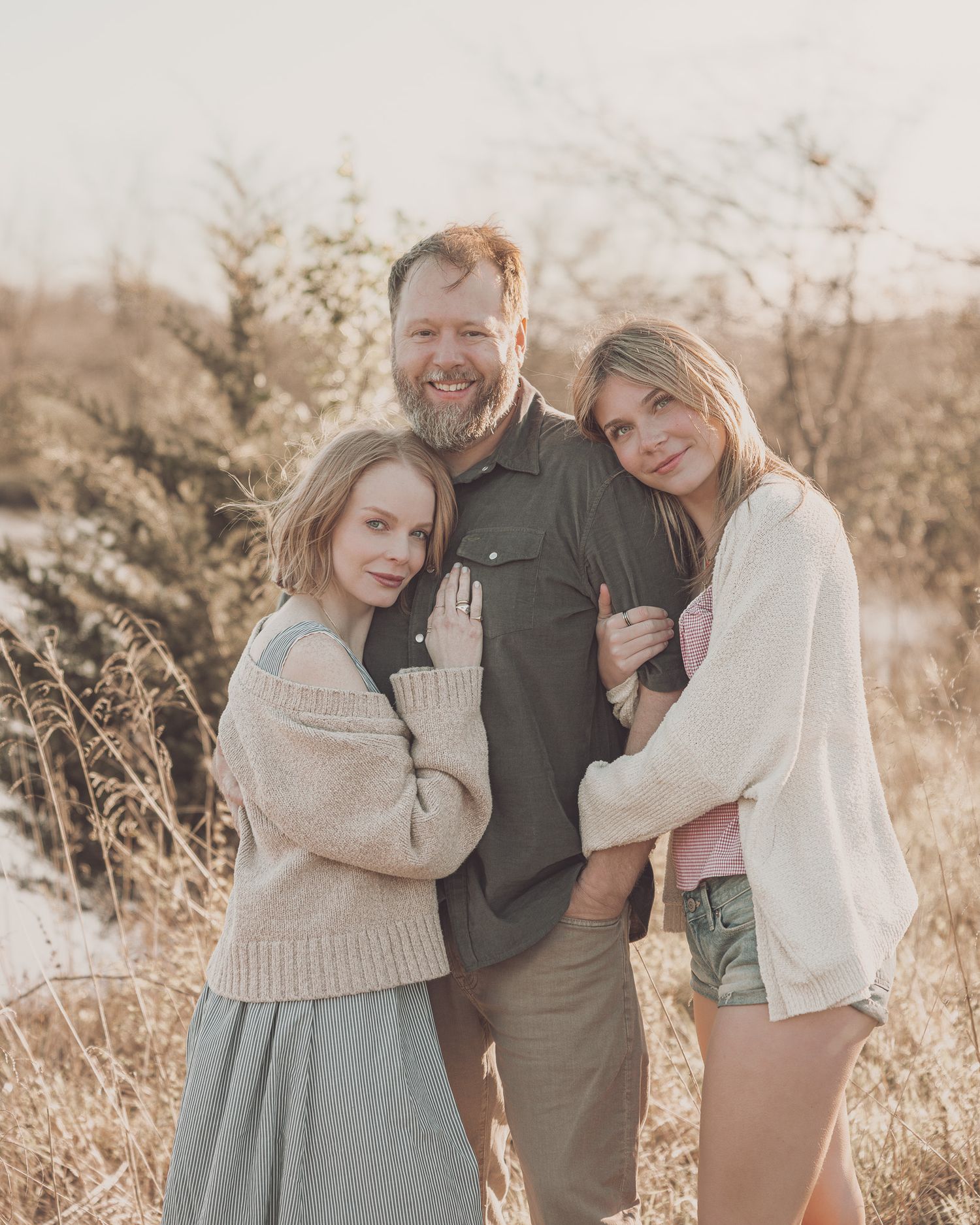 A bearded man poses outdoors with two young women in a warm, golden field setting during fall.