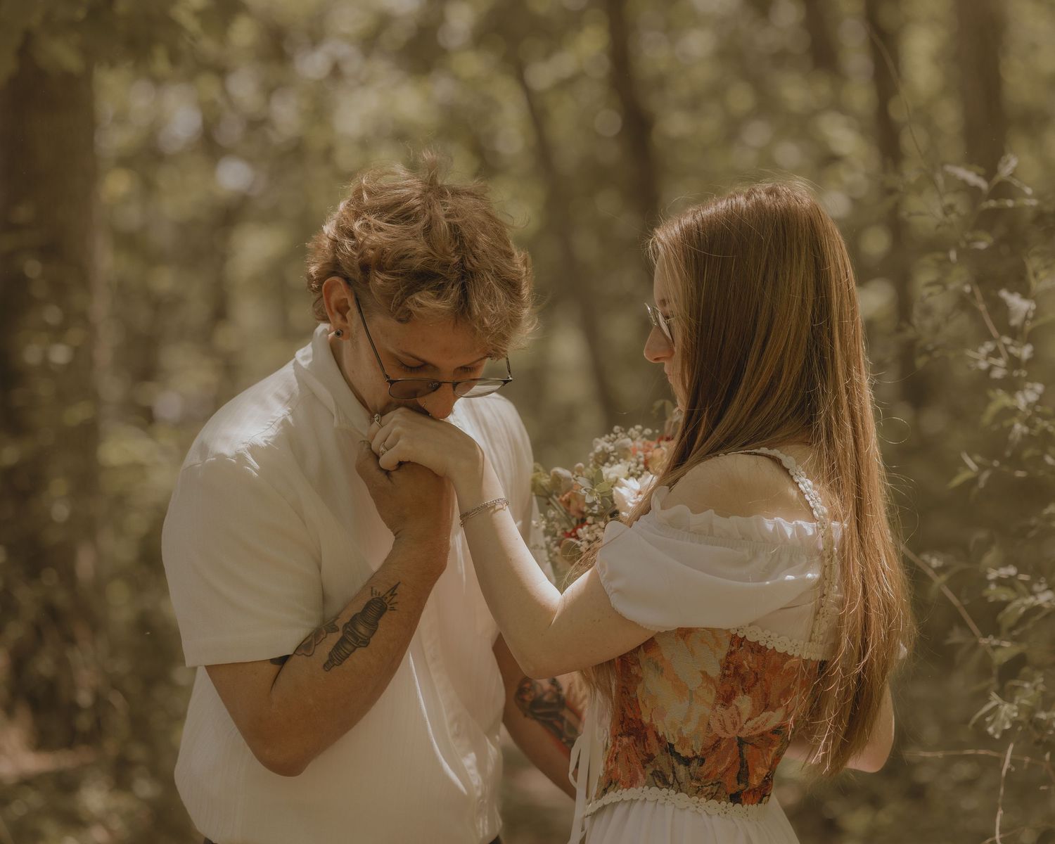 A romantic couple in a sunlit forest, man kissing woman's hand while she holds a wildflower bouquet.