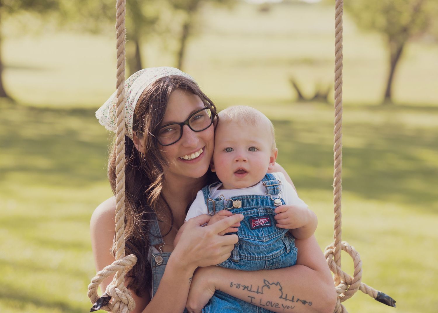 Smiling mother holding baby boy on wooden swing in sunny outdoor park setting.