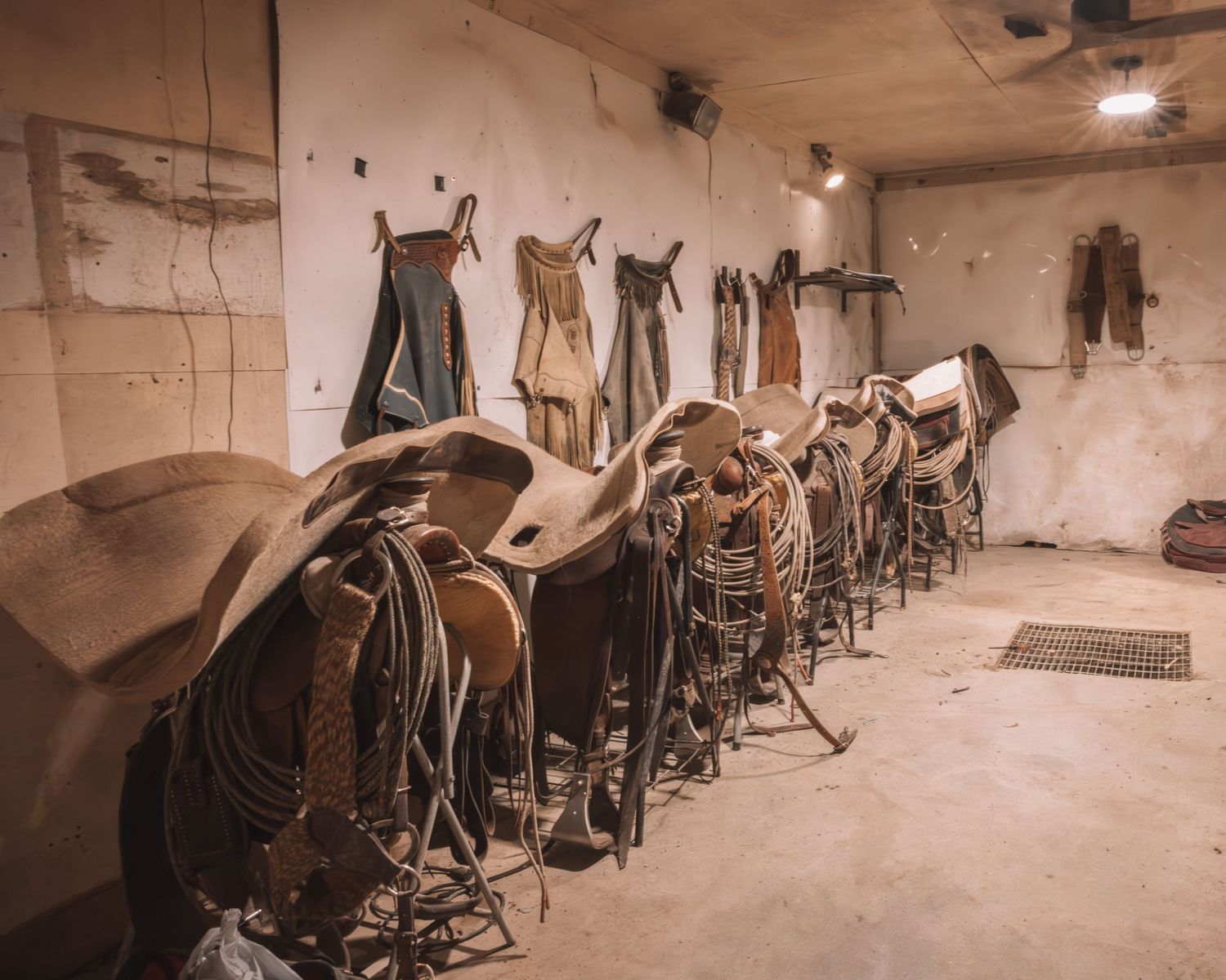 A rustic tack room displays rows of Western saddles and riding gear on a stable wall.