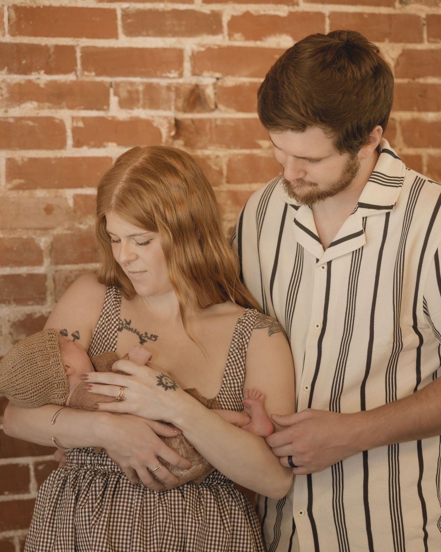 New parents holding newborn baby together in front of rustic brick wall in studio portrait.