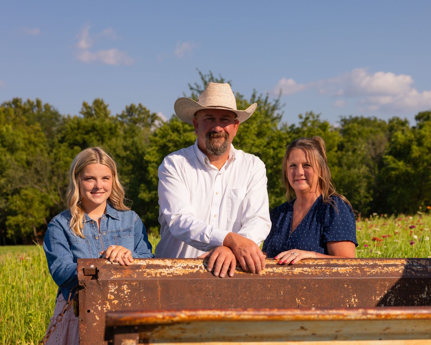 A family of three poses outdoors by a rustic farm truck in a green field on a sunny day.