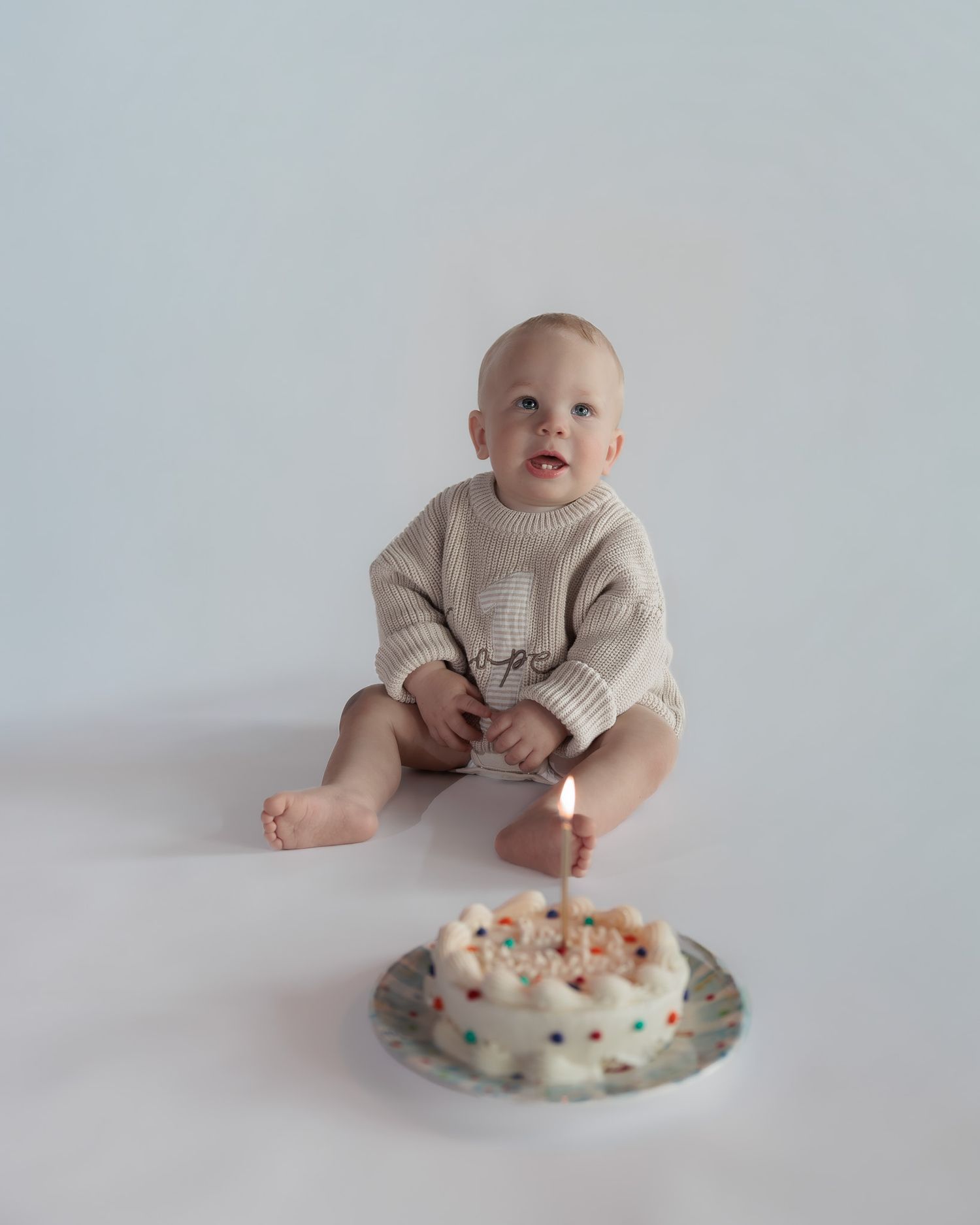 Baby boy in beige knit romper sits before a small white birthday cake with one lit candle on a white background.