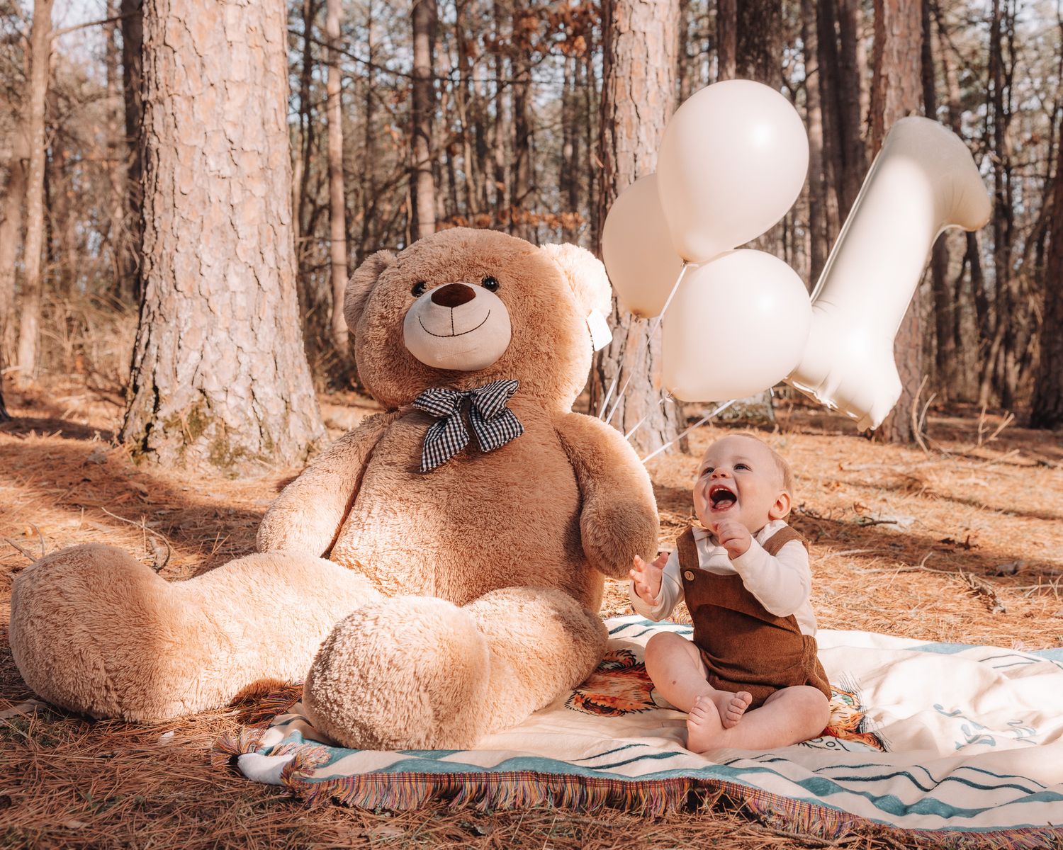 A cozy outdoor family photoshoot setup in a forest with a large teddy bear and blanket on the ground.
