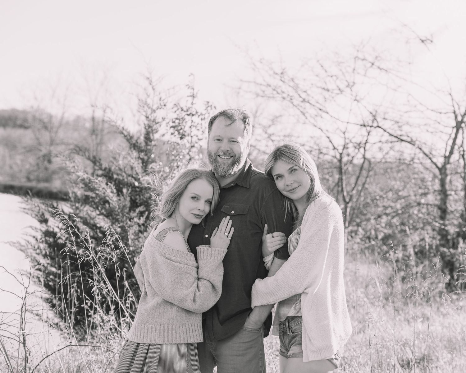 Black and white family portrait of a bearded man with two girls hugging him outdoors in a natural setting.