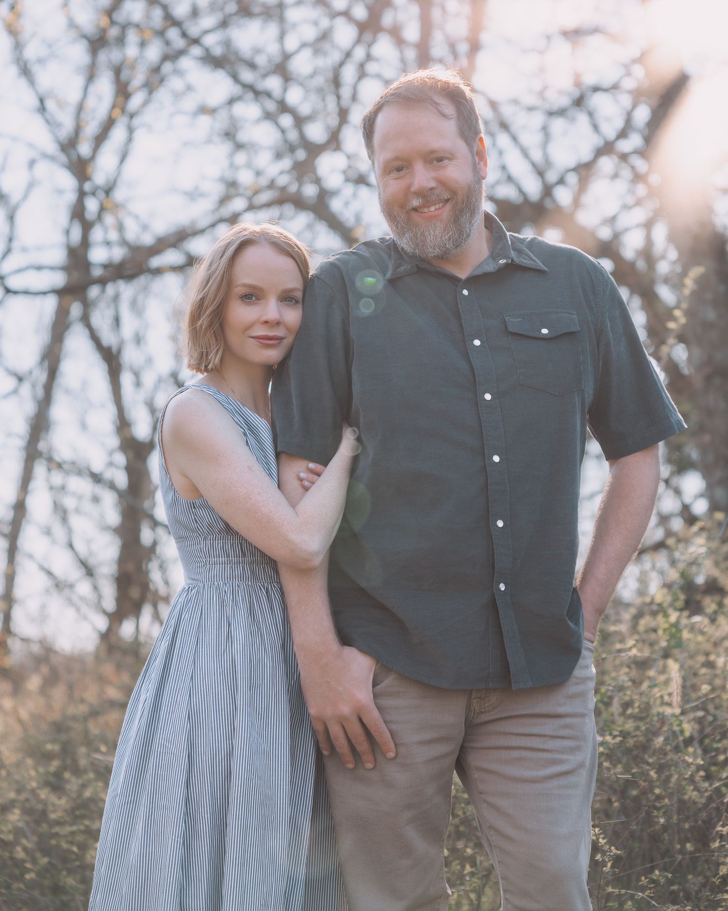 A smiling couple poses outdoors among trees at golden hour, woman in striped dress, man in gray shirt.