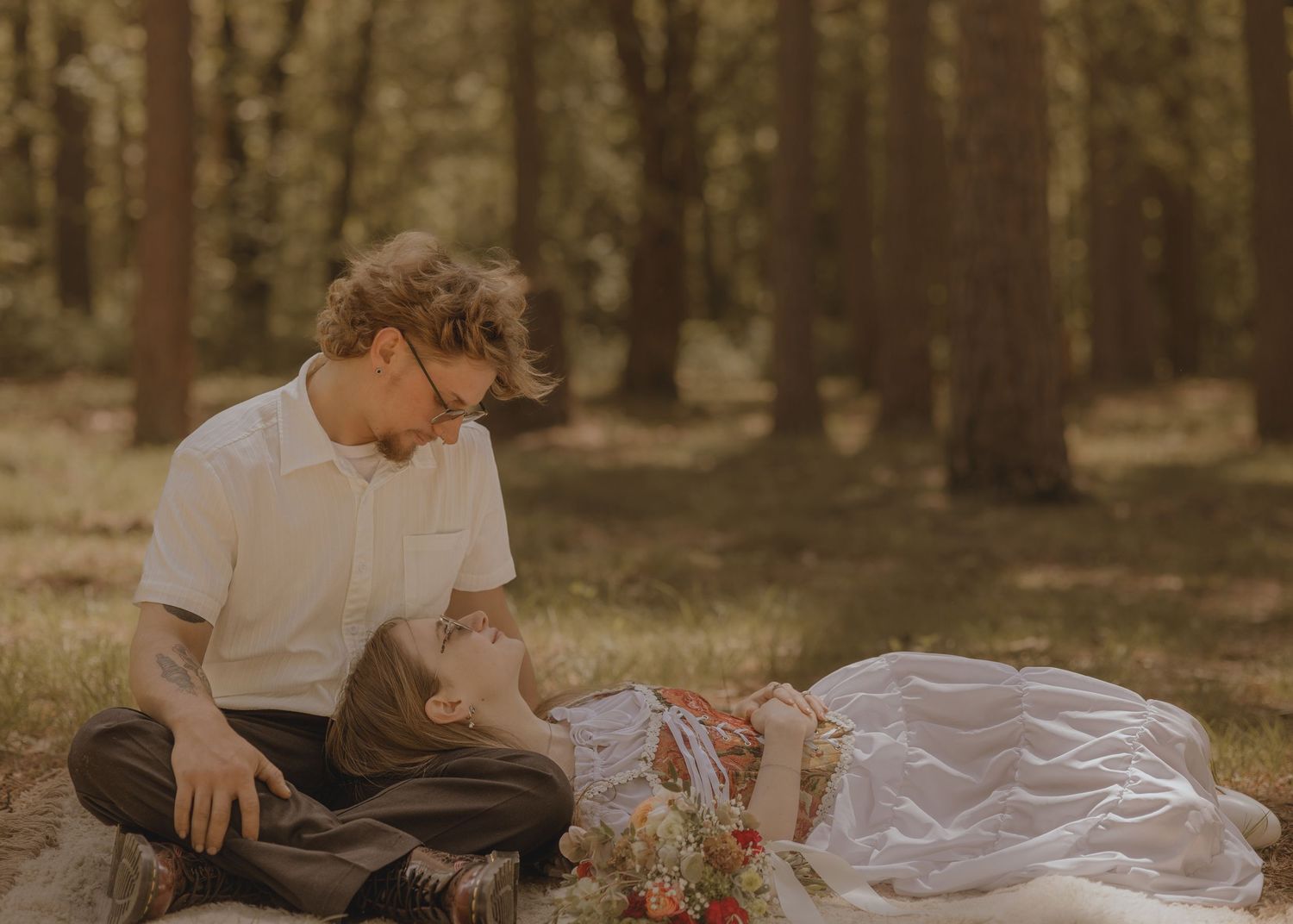 Romantic couple sharing a tender moment on a picnic blanket in a sunlit forest, with flowers nearby.