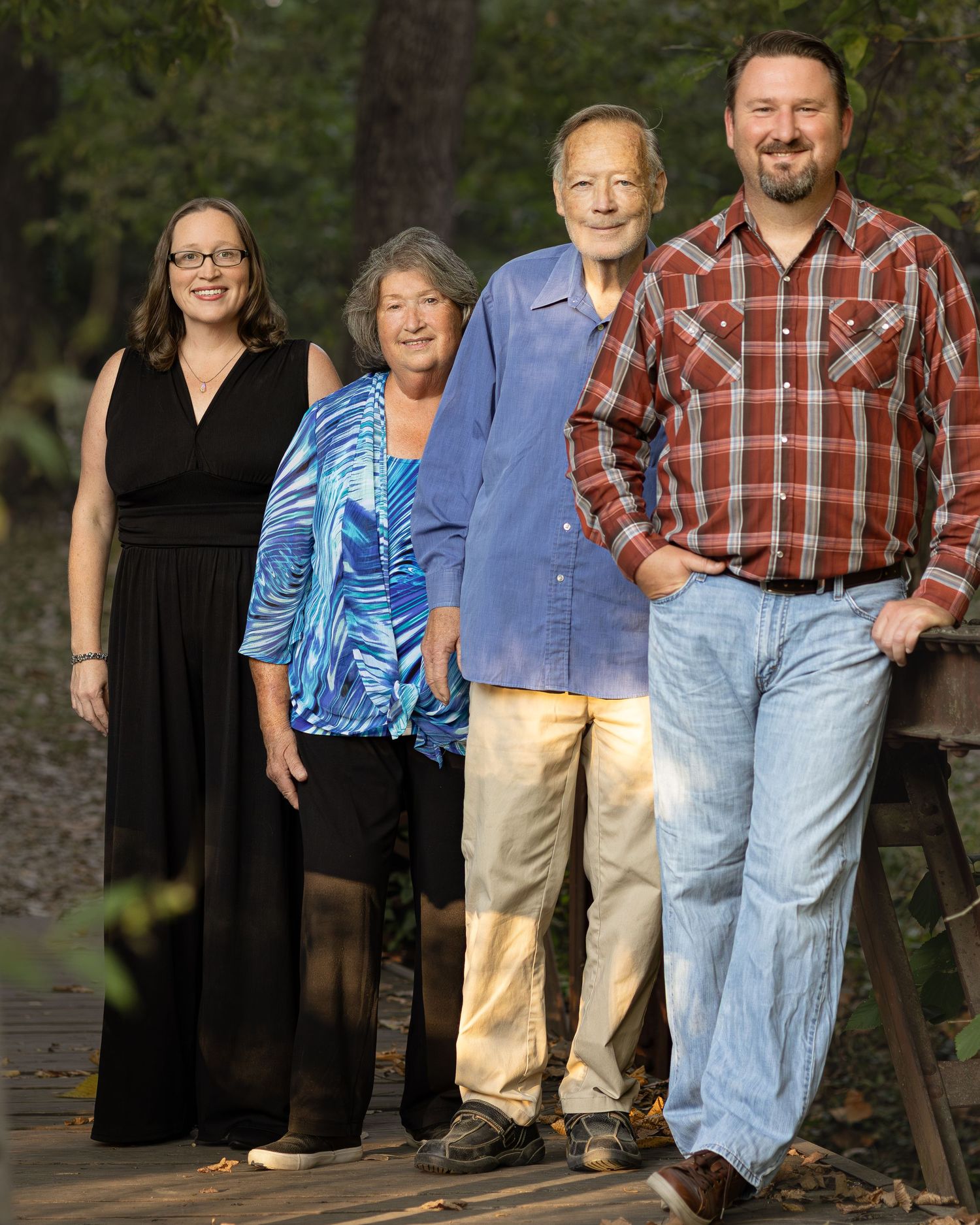 Four adults smiling together on a wooden bridge surrounded by lush green trees in an outdoor family portrait.