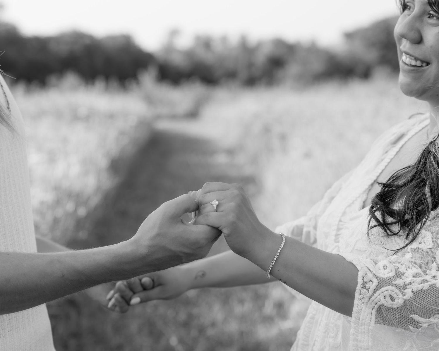 Black and white photo of couple holding hands outdoors, woman smiling in lace dress during engagement session.