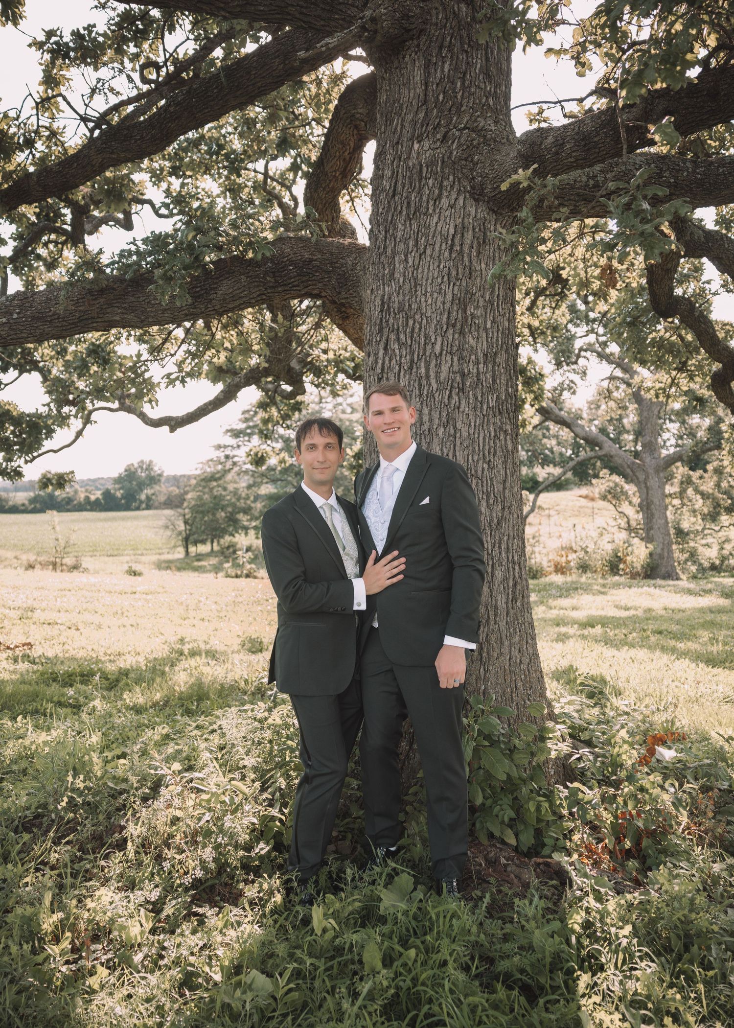 Two grooms in dark suits pose together leaning against a large oak tree in a scenic outdoor setting.