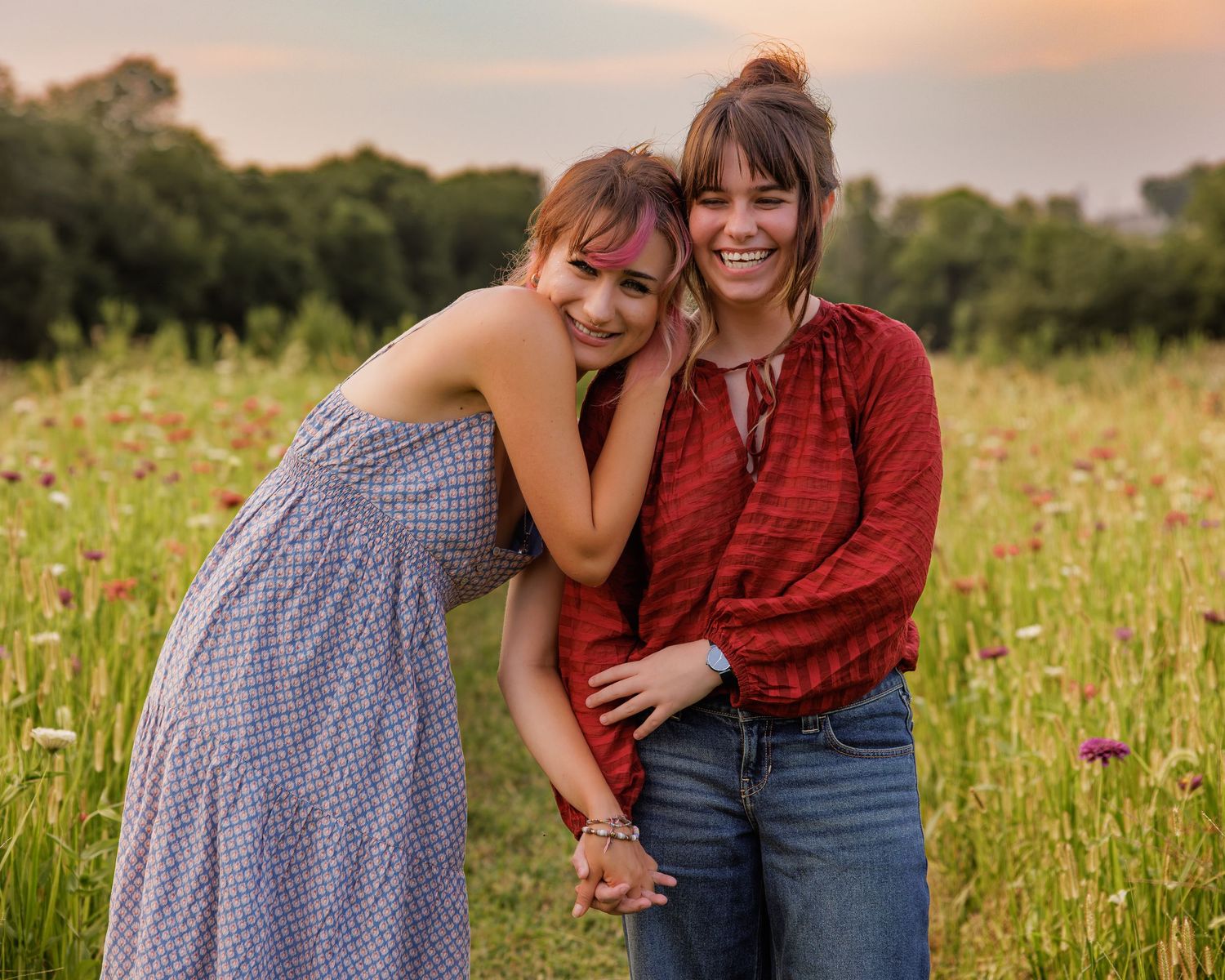 Two women smiling and embracing in a wildflower field at sunset, one in a blue dress, one in a red blouse.