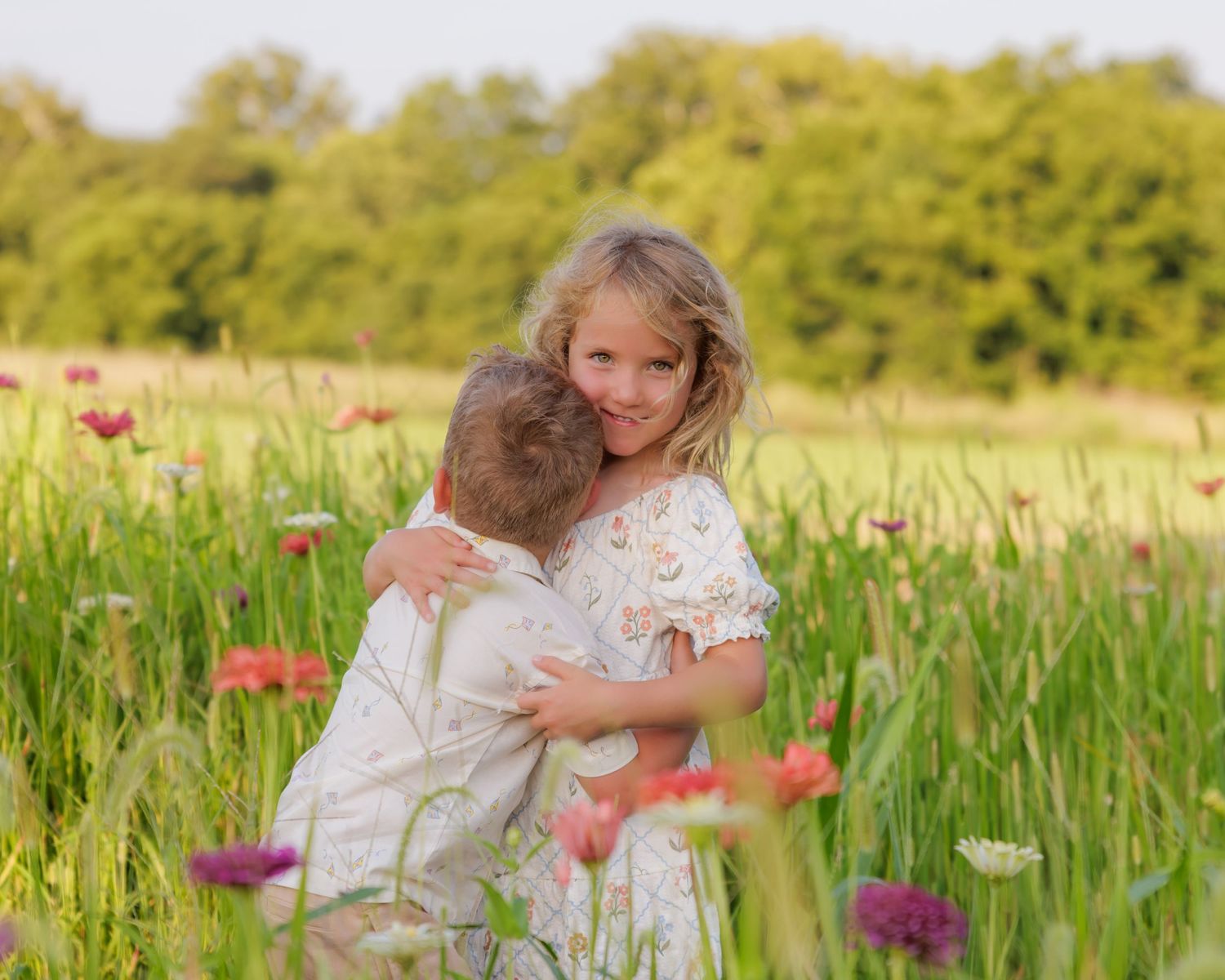 Two children hugging in a colorful wildflower meadow, surrounded by pink and purple blooms with green trees in background.