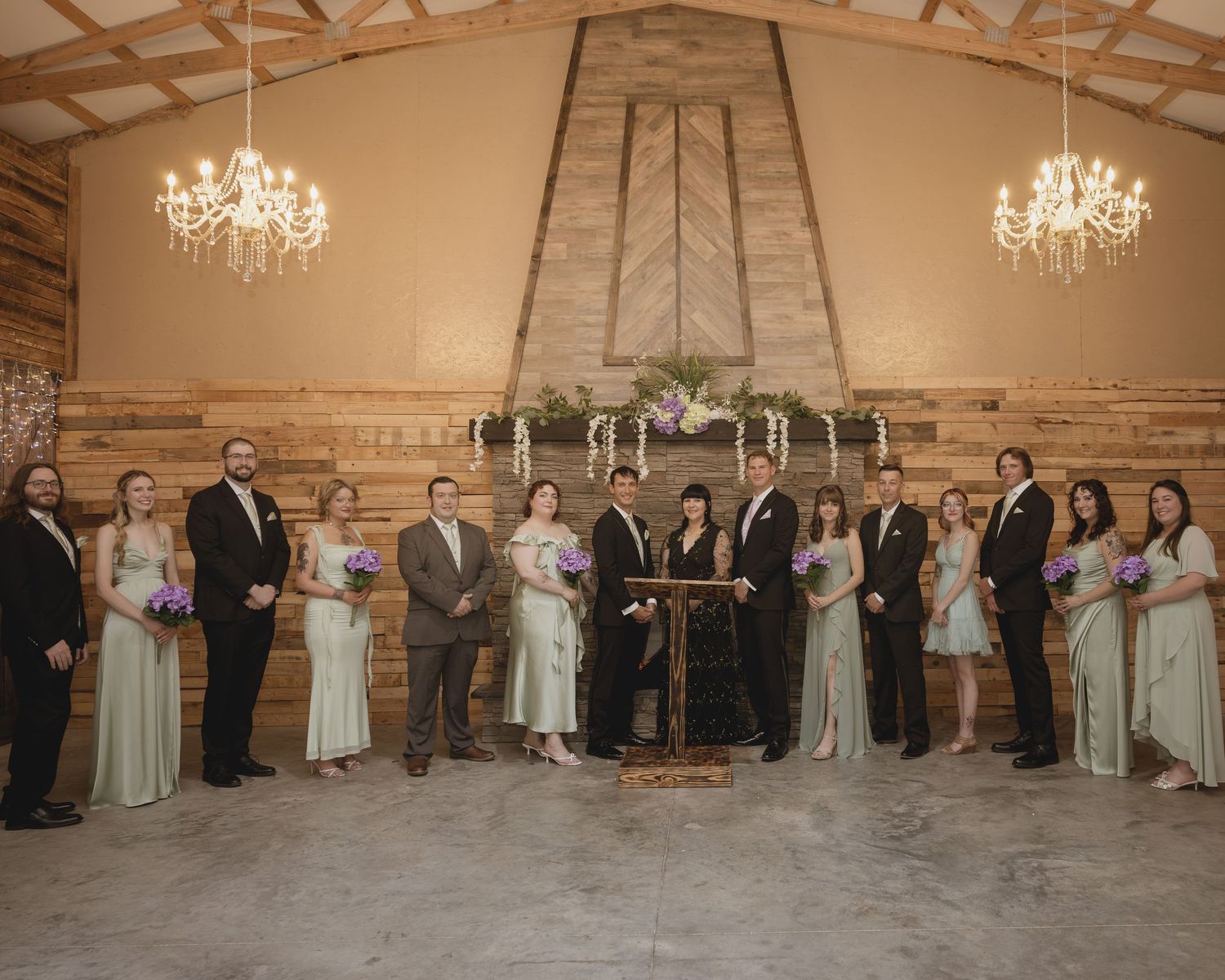 Wedding party poses in rustic barn venue with chandeliers and fireplace backdrop during indoor ceremony.