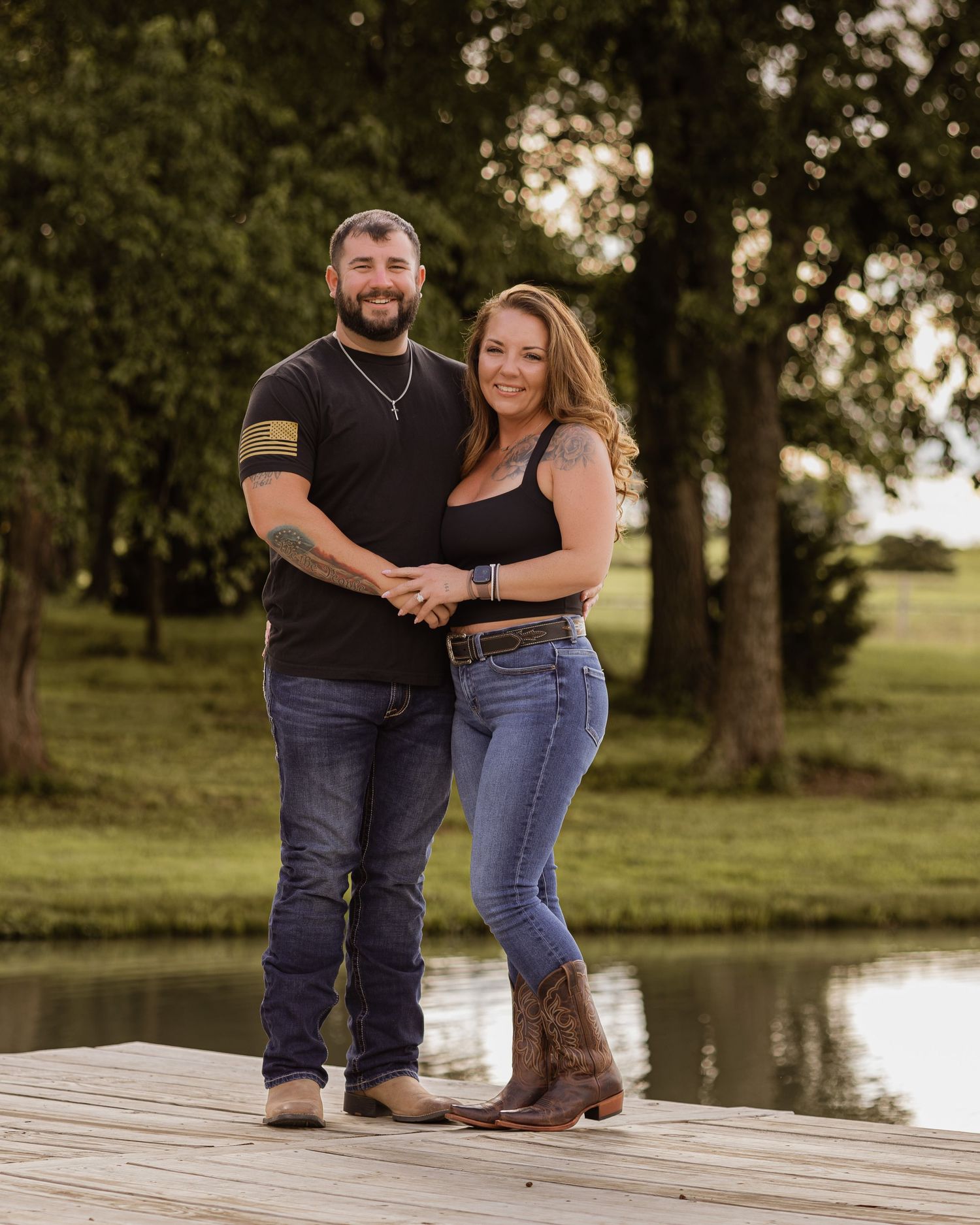 Couple smiling together on a wooden dock by a pond surrounded by lush green trees.