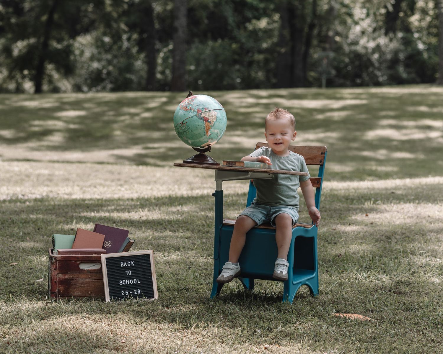 A small child sits in a blue chair next to a globe and vintage suitcases on a grassy lawn.