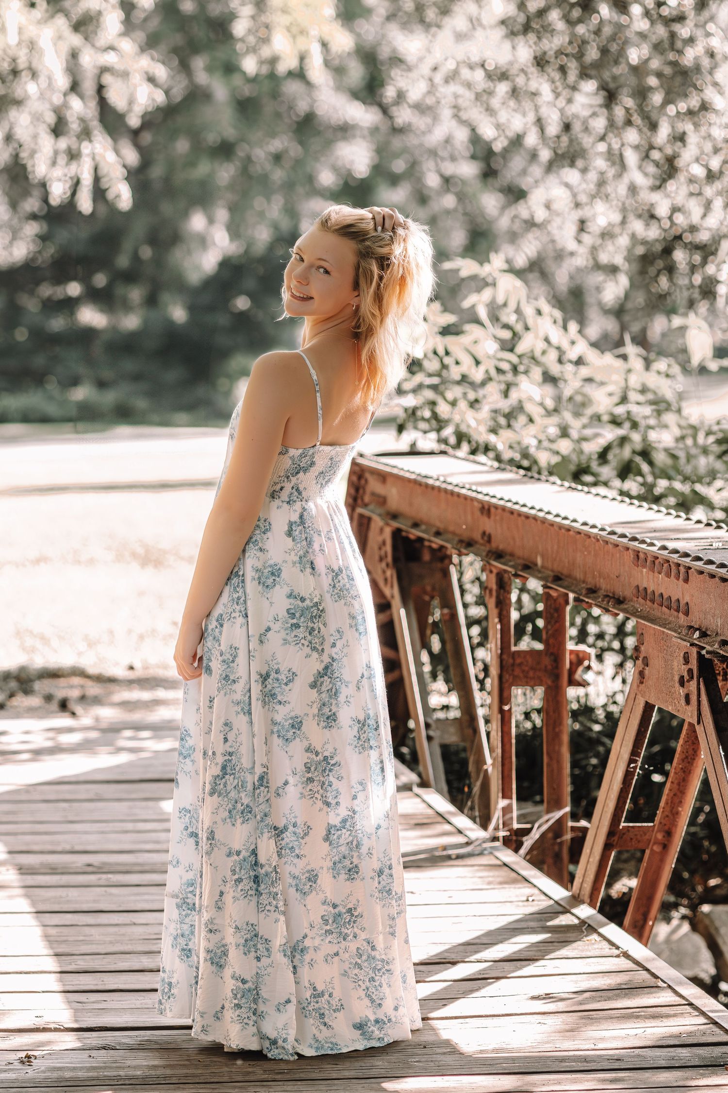 A person in a floral white dress sits on a rustic wooden bridge in soft sunlit surroundings.