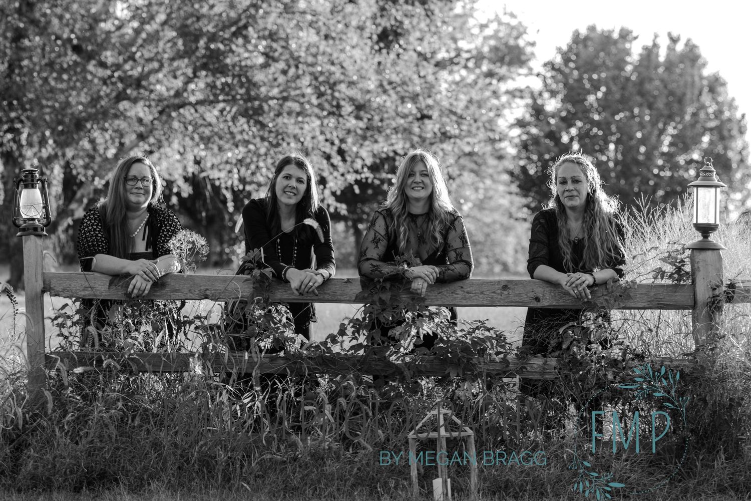 Group of people sitting on a rustic wooden fence in a pastoral outdoor setting with trees in the background.