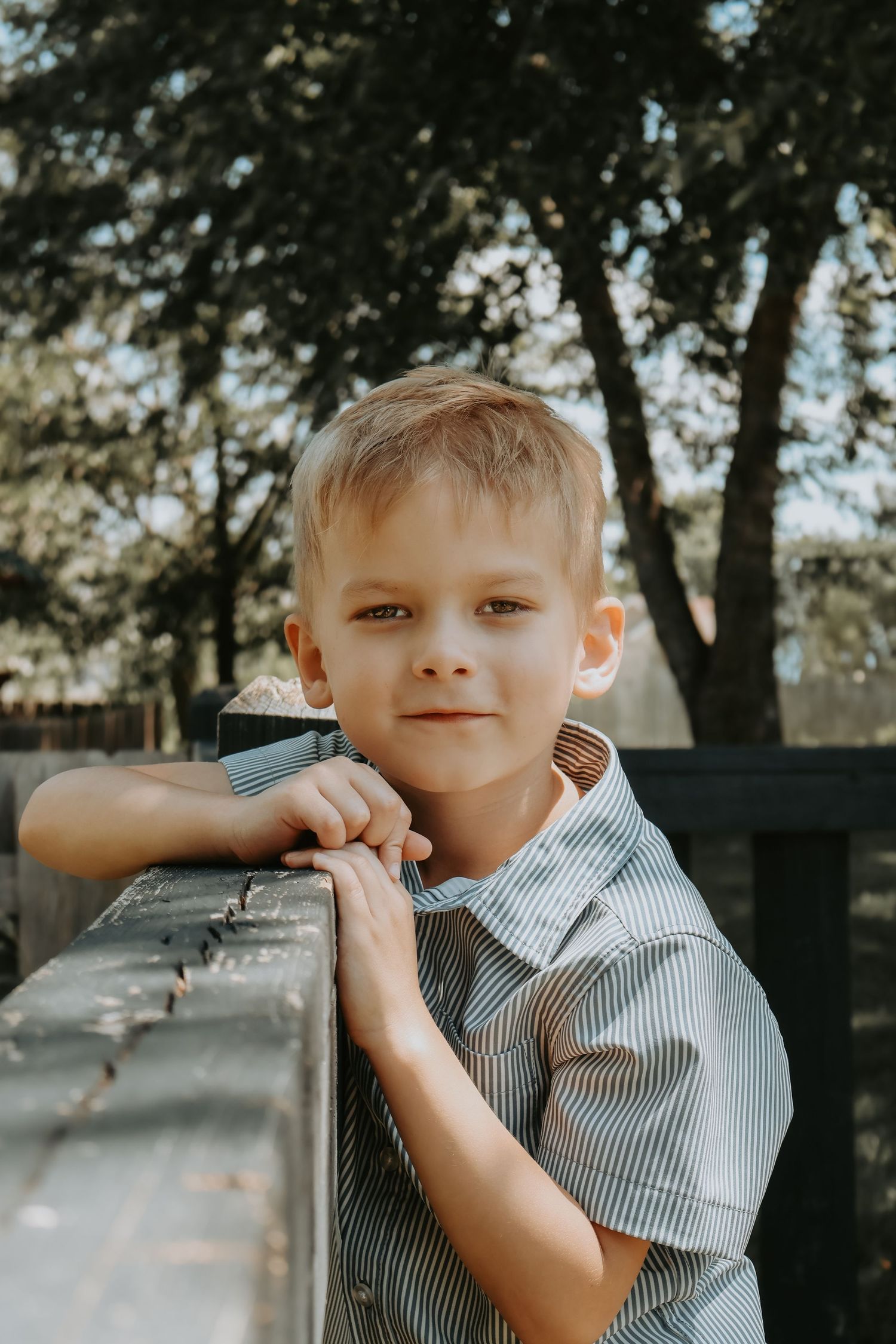A young child in a striped button-down shirt leans against a wooden railing outdoors with trees in the background.