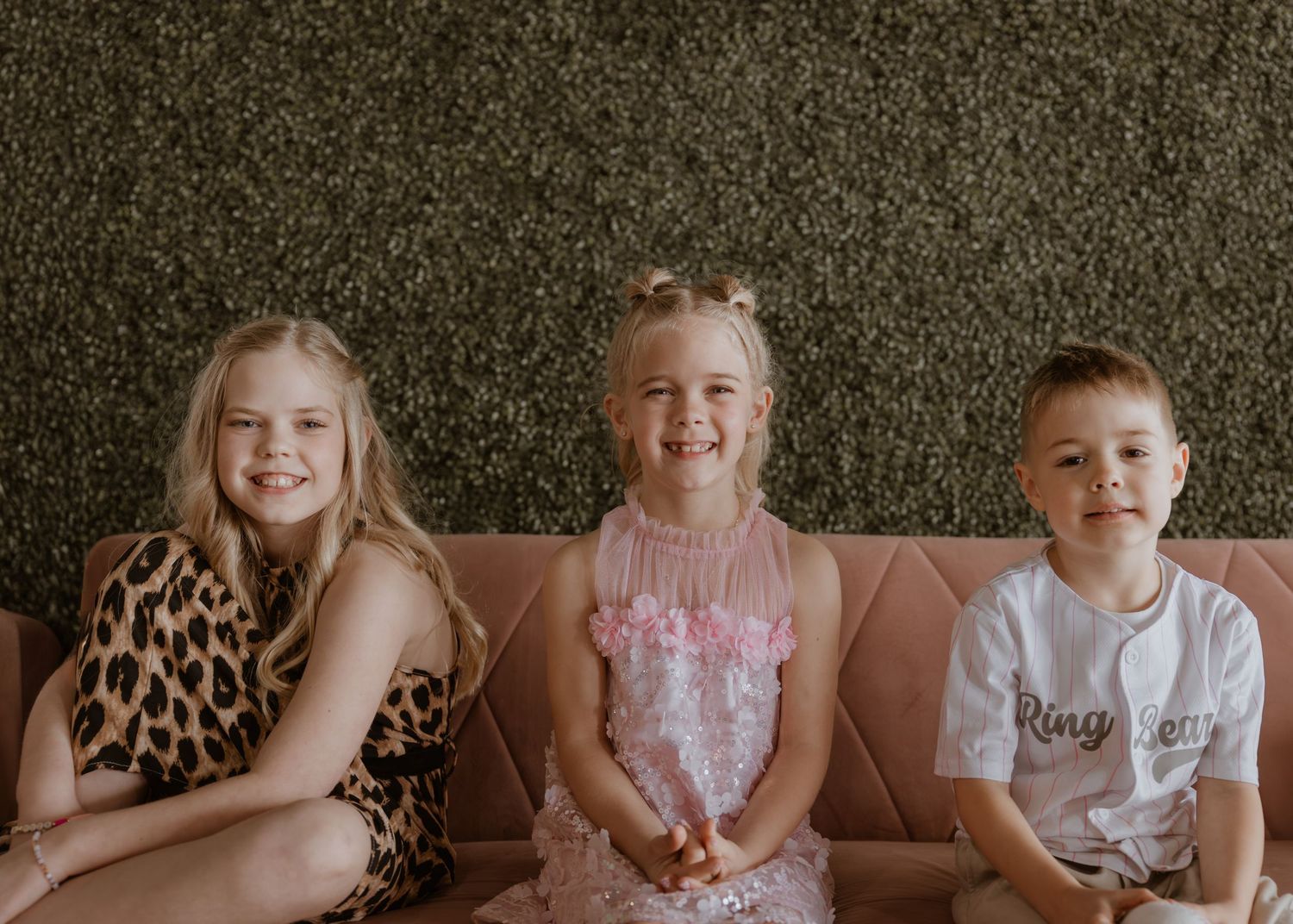 Three smiling children in festive attire sitting on pink velvet couch against green hedge wall backdrop.