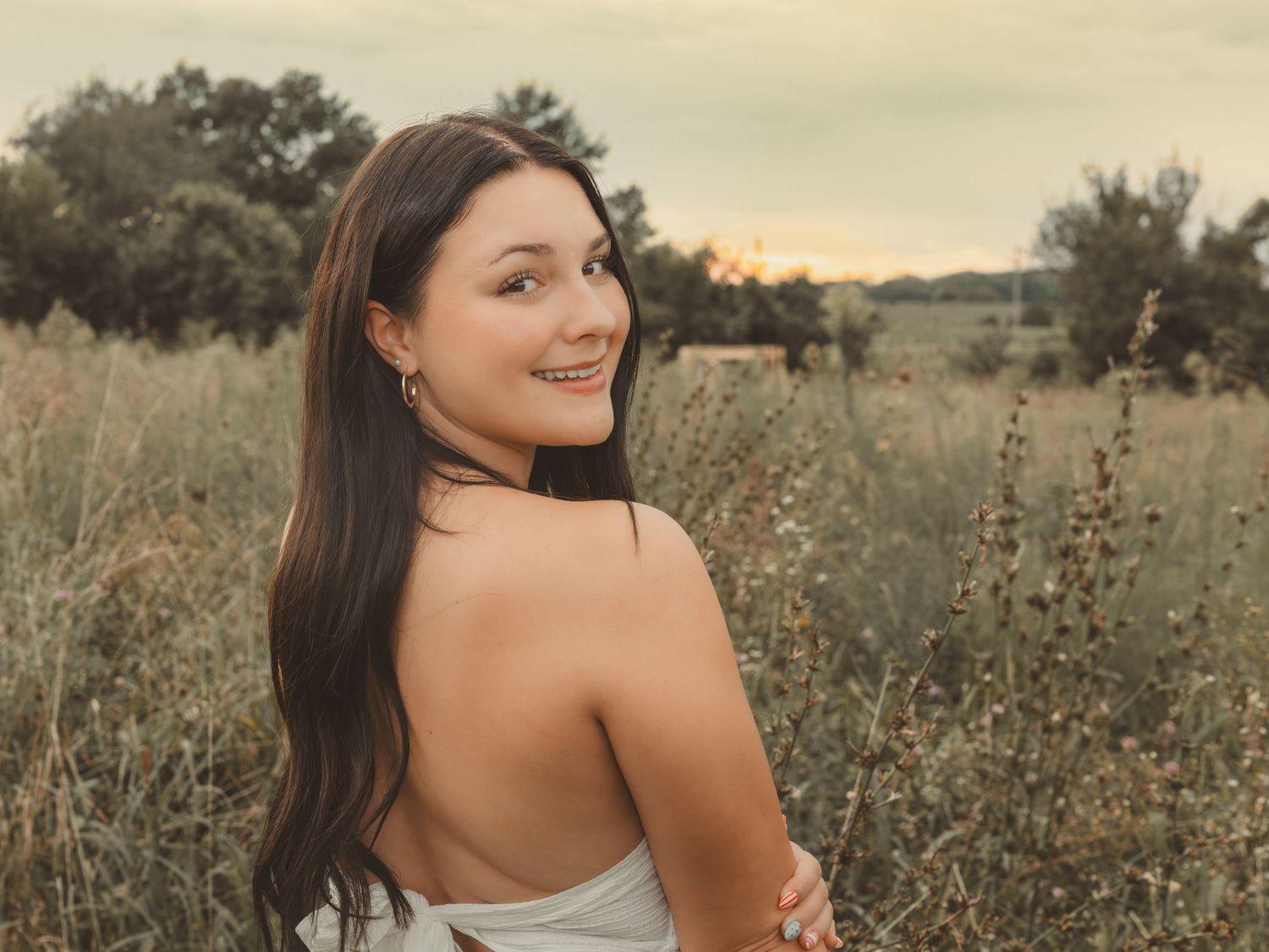 Young woman with long dark hair smiling over her shoulder in a golden field at sunset.