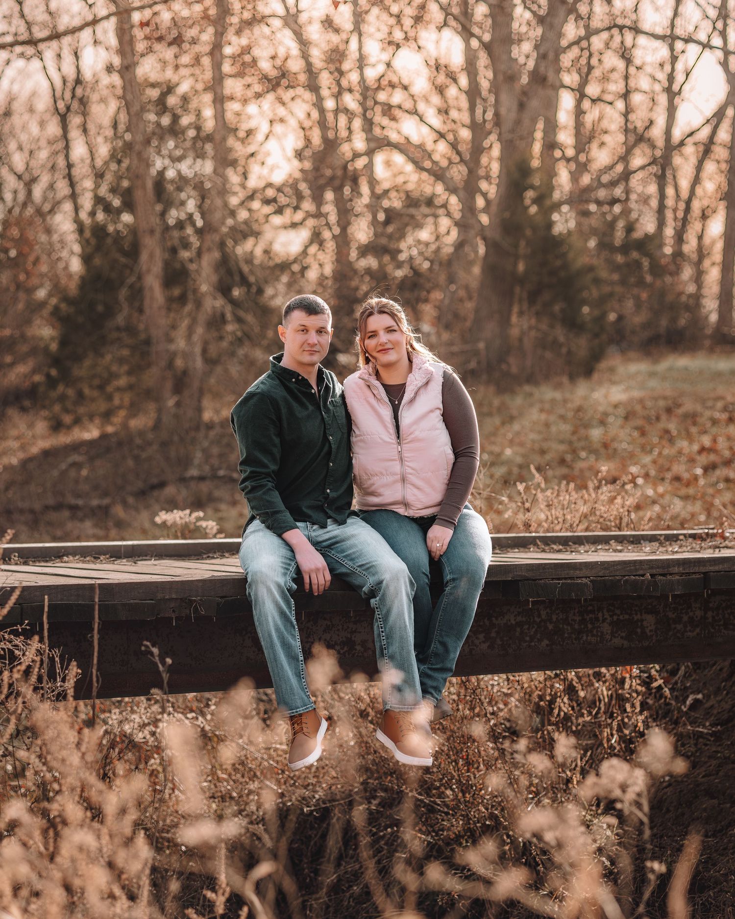 A couple sits together on a rustic wooden fence surrounded by autumn foliage at sunset.