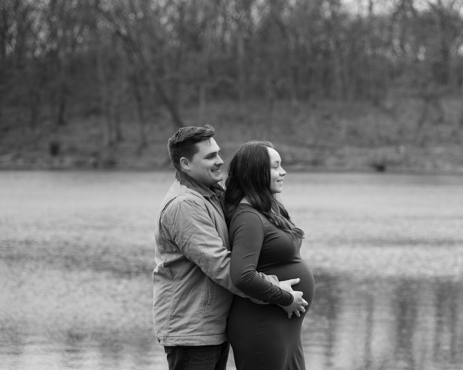 A couple embraces by a serene lake in black and white, with the woman cradling her pregnant belly.