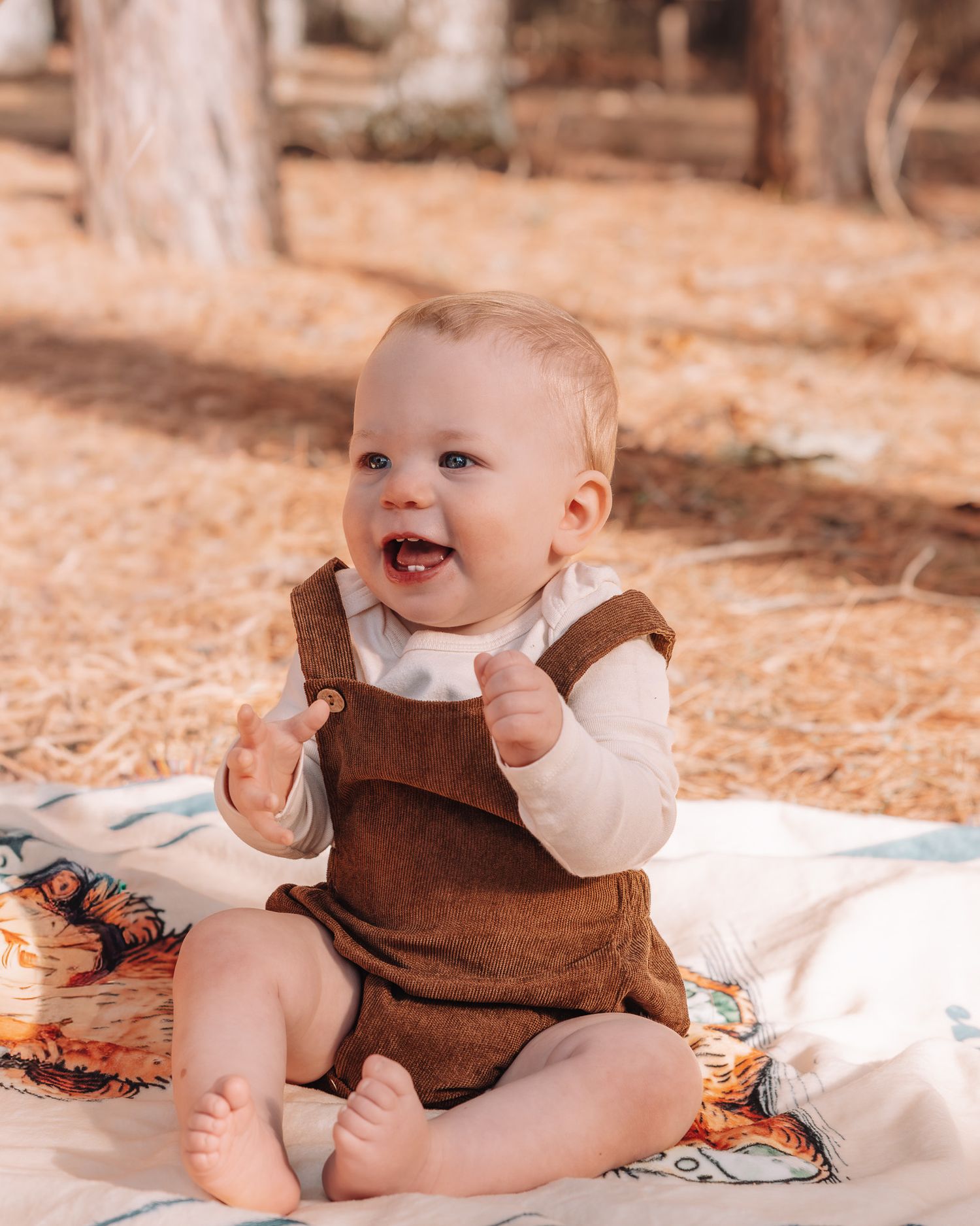Happy baby wearing brown overalls and white long sleeve shirt sits on blanket outdoors in autumn setting.