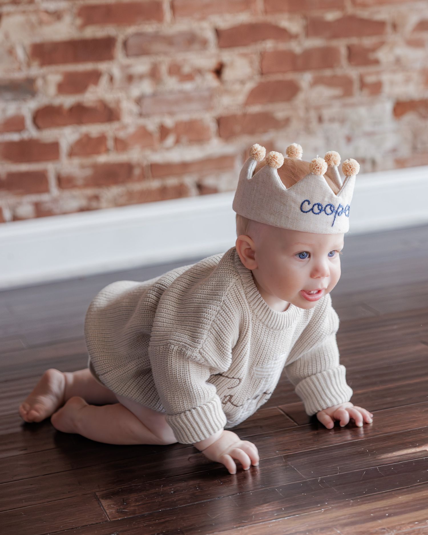 Adorable baby boy Cooper crawls on hardwood floor wearing a personalized fabric crown hat against a brick wall backdrop.