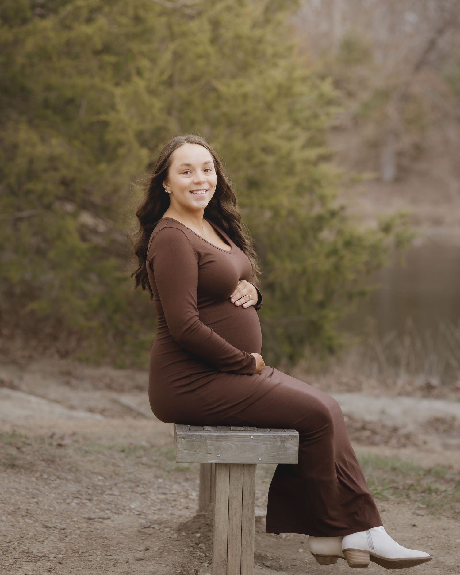 A person in a brown dress poses on a wooden bench against a scenic natural backdrop with greenery.