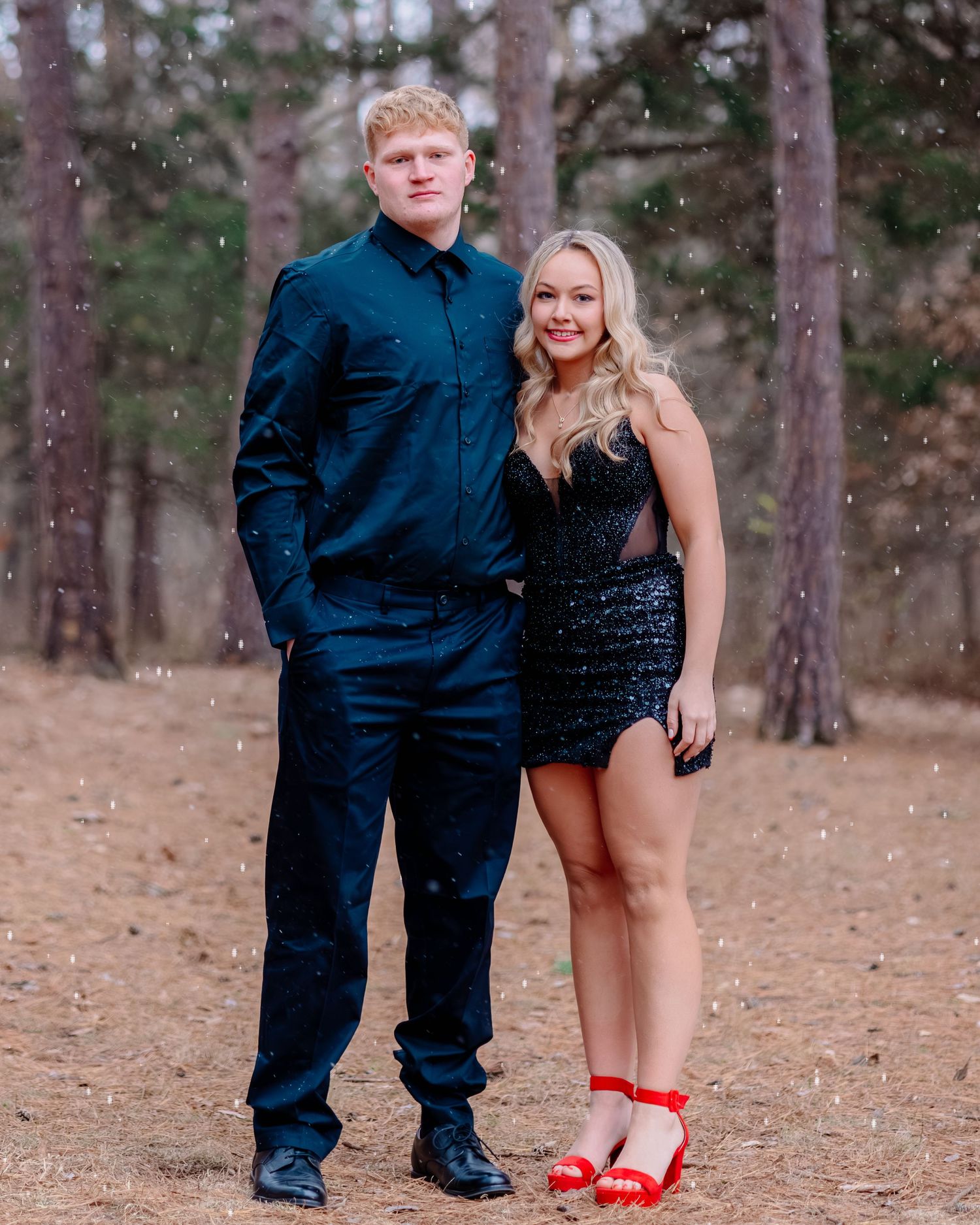 A couple in formal attire stands together in a wooded setting, with her wearing red shoes and him in navy blue.