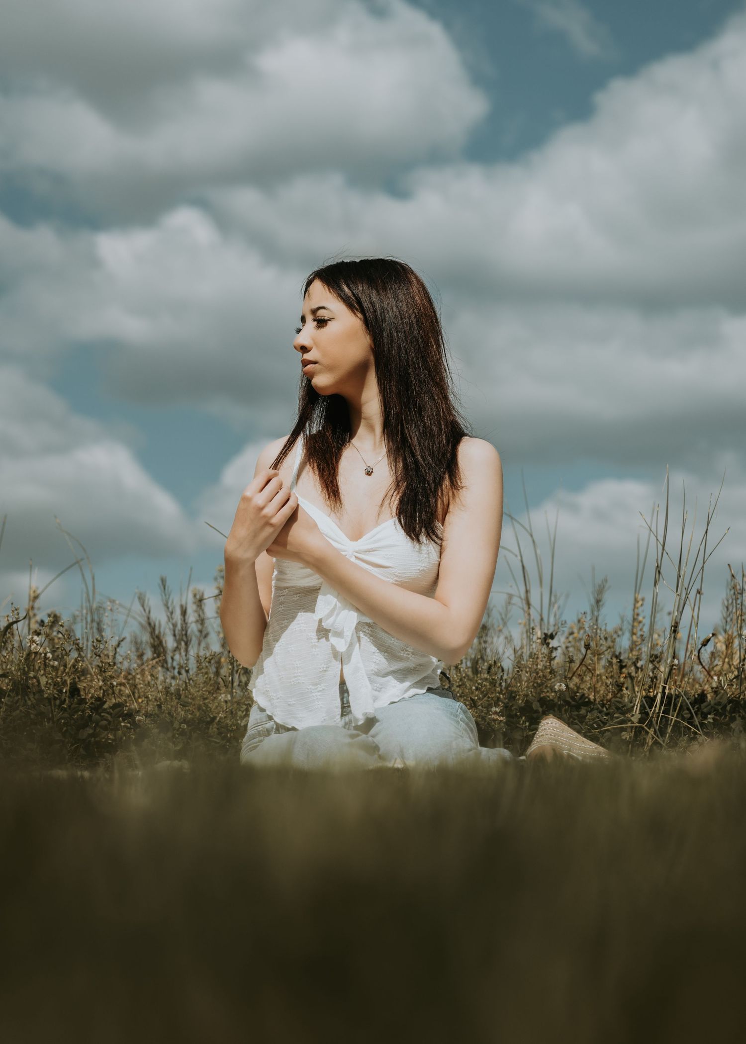 Young woman in white top sitting in a golden field of tall grass under a dramatic cloudy sky.