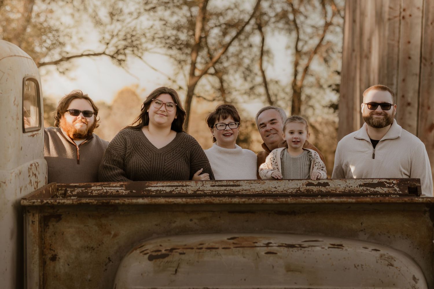 Family of six posing behind a rustic vintage truck bed outdoors in warm golden hour light.