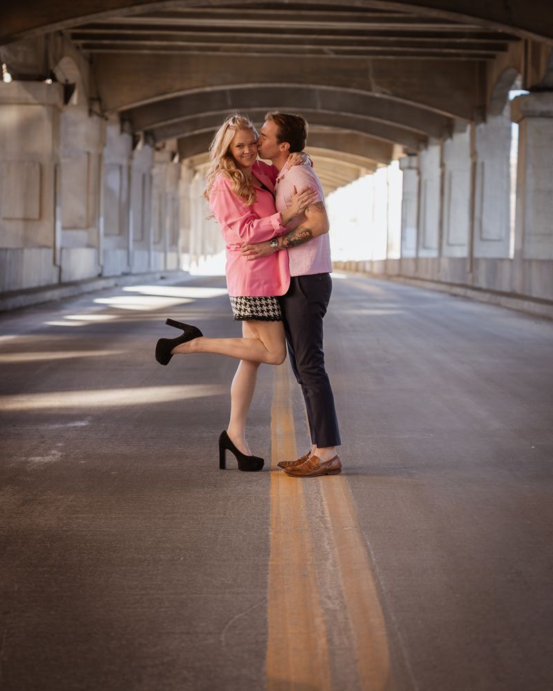 Couple embracing and laughing under a bridge overpass, woman in pink top lifting one heel in black high heels.