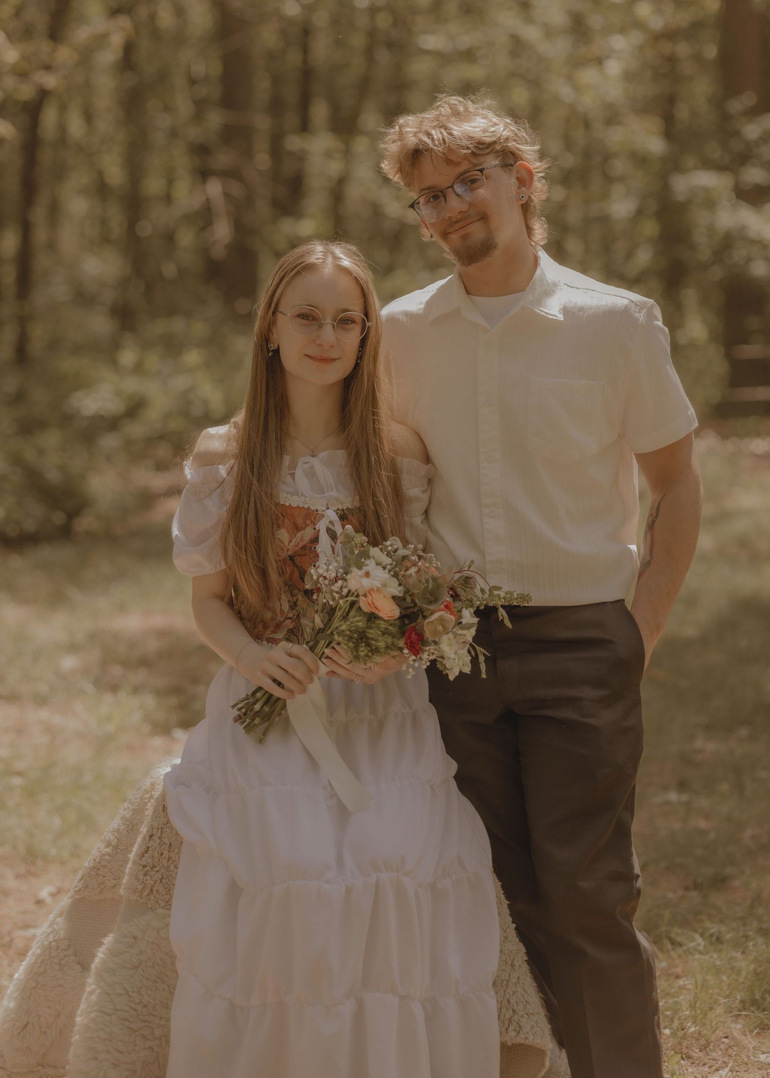 A couple poses in a forest, bride in white dress holding wildflower bouquet, groom in white shirt and dark pants.