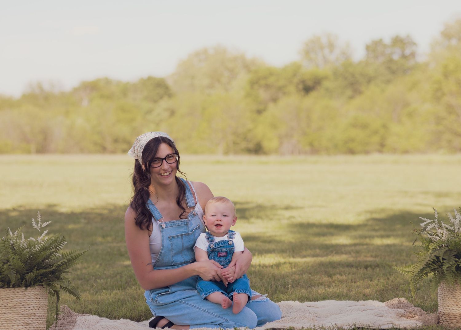A smiling woman in denim overalls sits on a blanket outdoors holding a baby in a sunny green field.