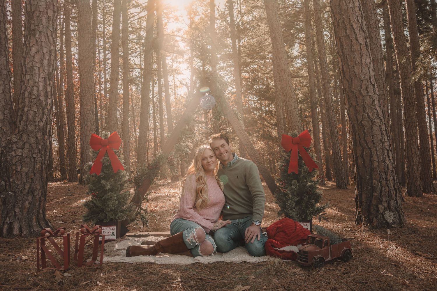 A romantic holiday scene in a forest with red Christmas decor, blanket, and sunlight streaming through tall trees.
