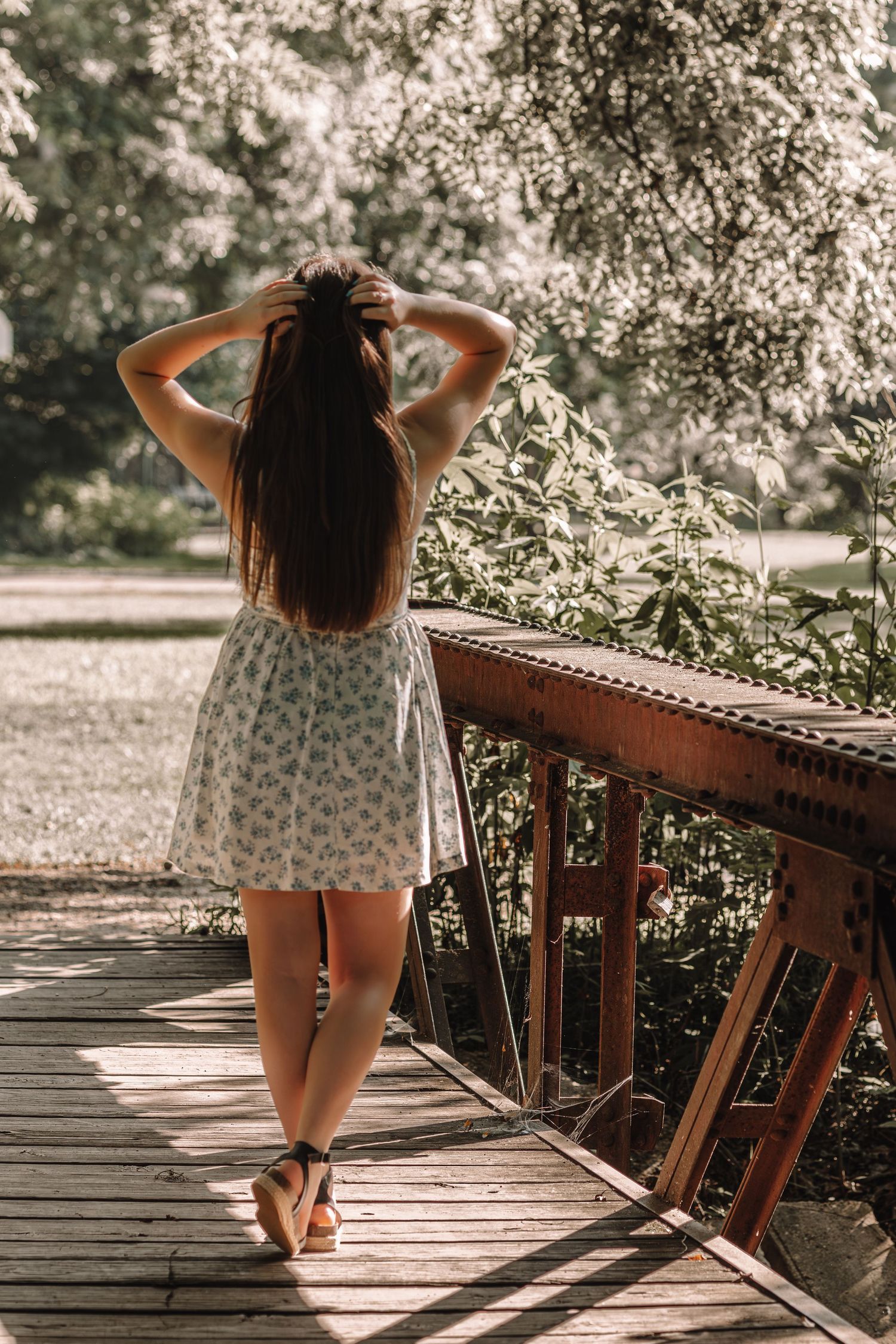 A person in a floral dress sits contemplatively on a wooden bridge railing during a sunny afternoon.