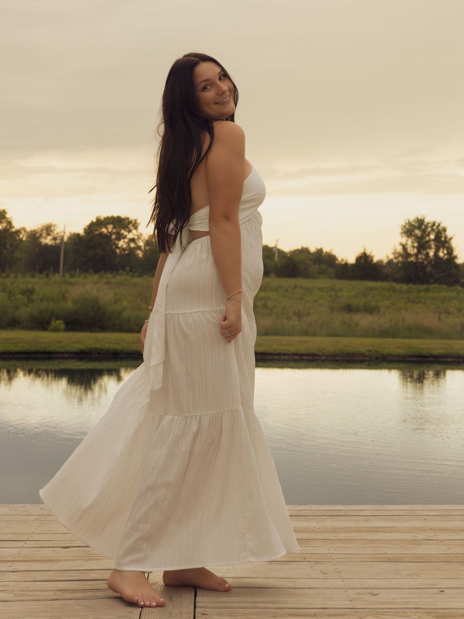 Brunette woman in white strapless crop top and maxi skirt on wooden dock at dusk.