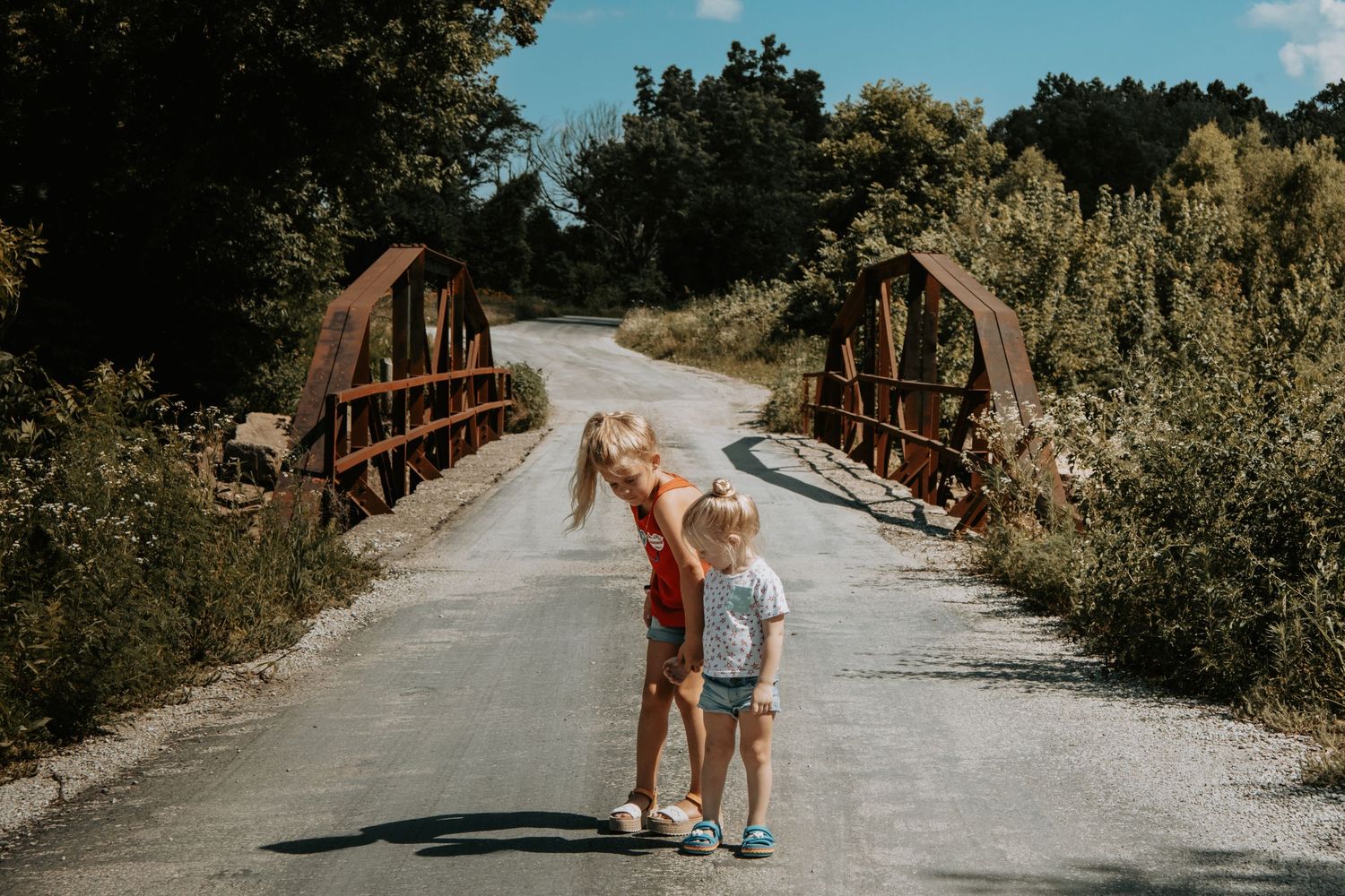 Two children stand on a curved wooden bridge surrounded by lush greenery on a sunny summer day.