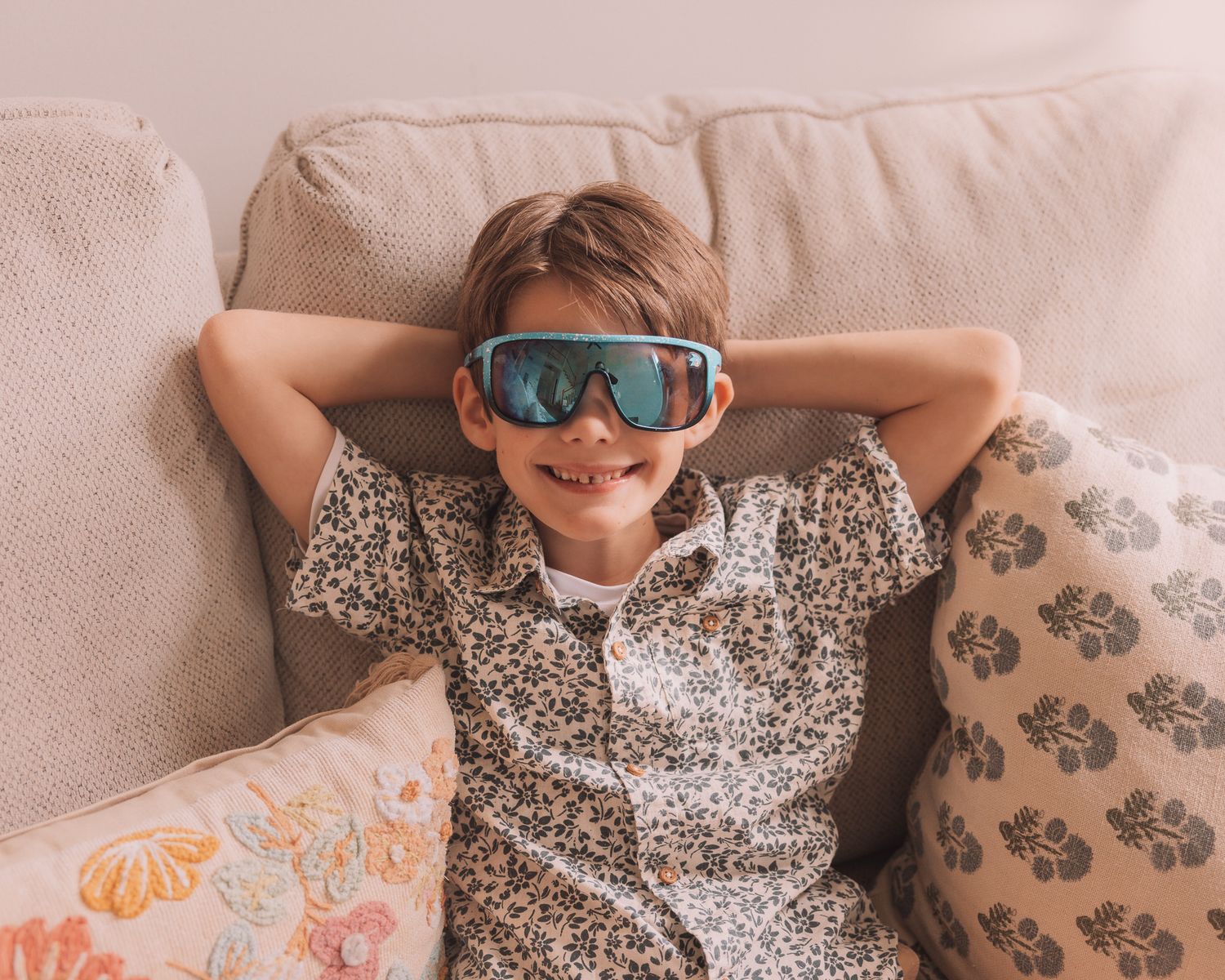 A cheerful boy in a leopard print shirt relaxes on a couch wearing oversized blue sunglasses.